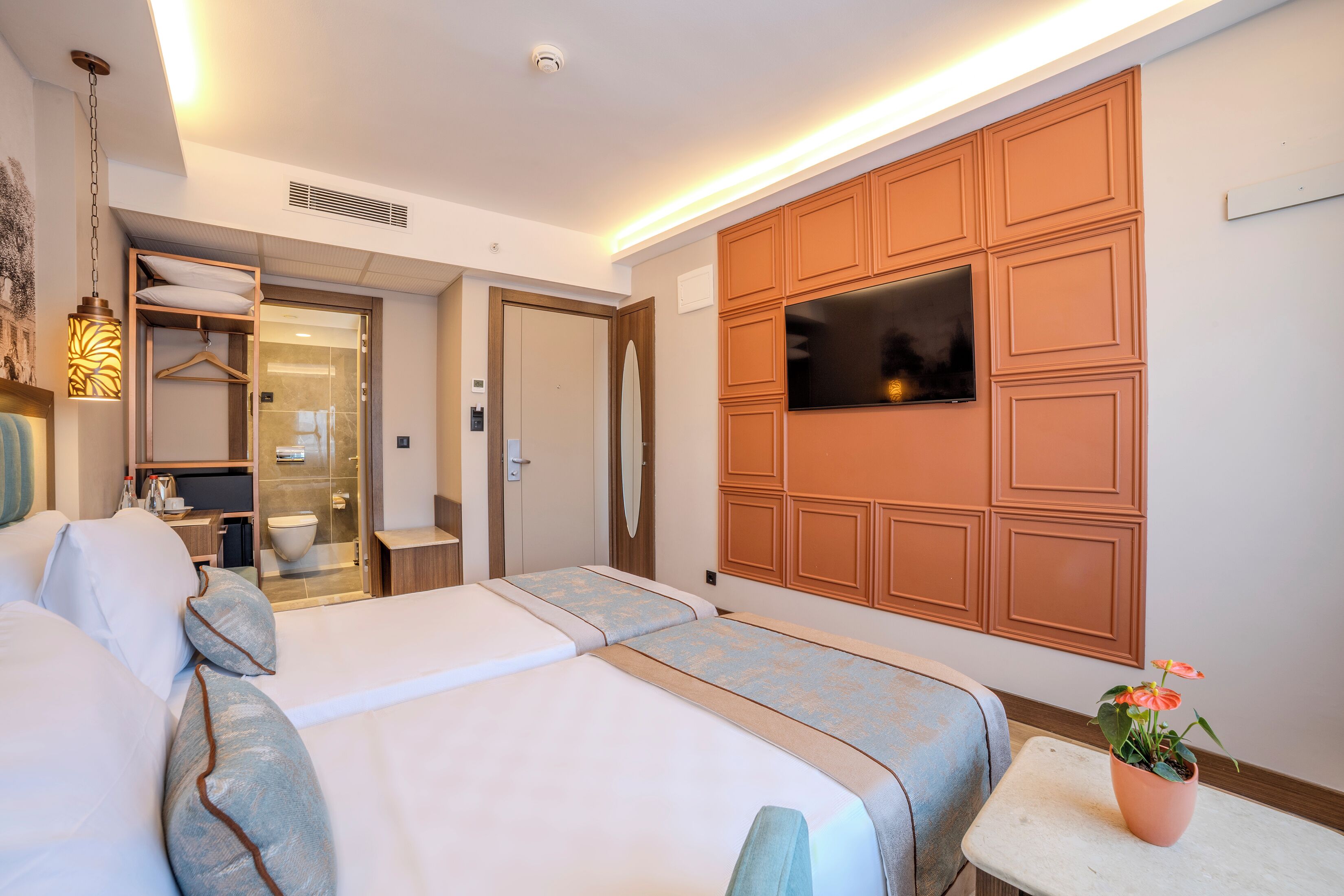 A hotel room with two beds, a TV mounted on the wall, a small table with a plant, and a bathroom visible through an open door.