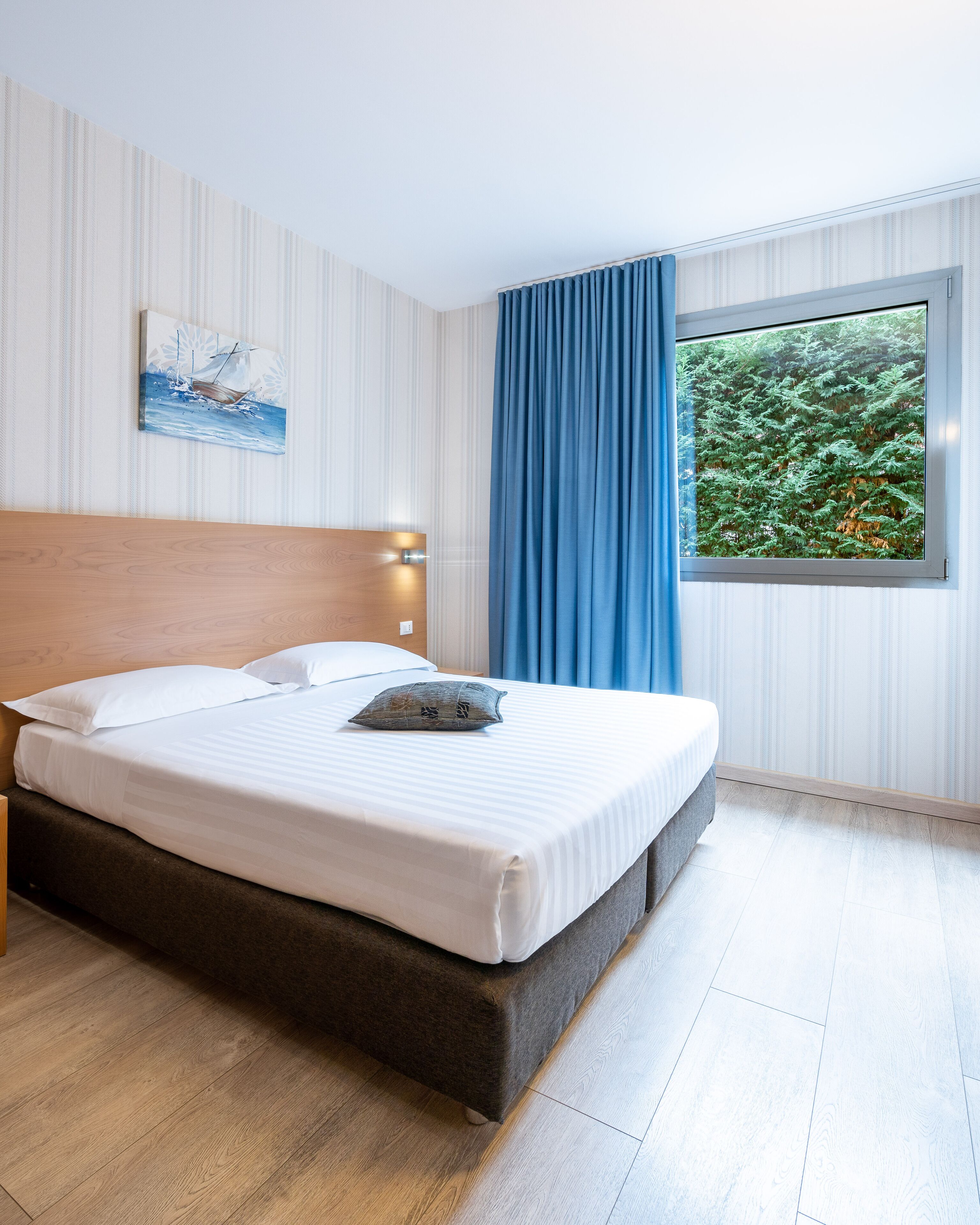 Una cama bien hecha con ropa blanca y un cabecero oscuro, un cuadro de un velero en la pared, y una ventana con cortina azul que da a un bosque.
