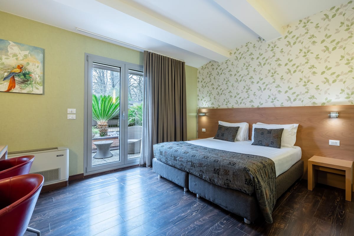 Une chambre d’hôtel avec un grand lit, une petite table, une chaise, une fenêtre donnant sur un espace verdoyant et un tableau au mur.