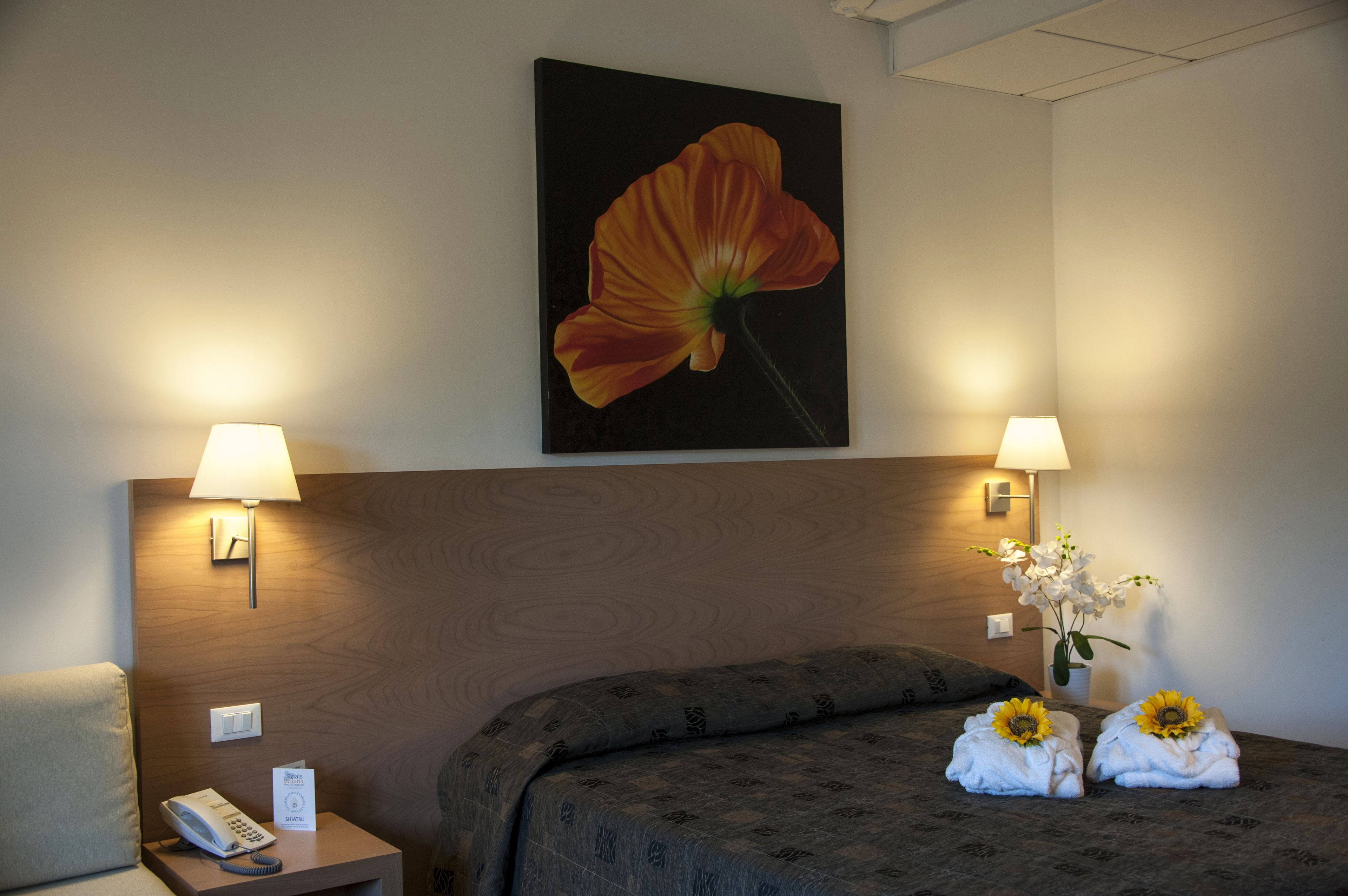 Habitación de hotel con cabecera de madera, dos lámparas de noche, un teléfono, un cartel y dos toallas con girasoles.