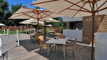 Een terras buiten met tafels en stoelen onder grote parasols, een houten terras en een modern gebouw op de achtergrond.