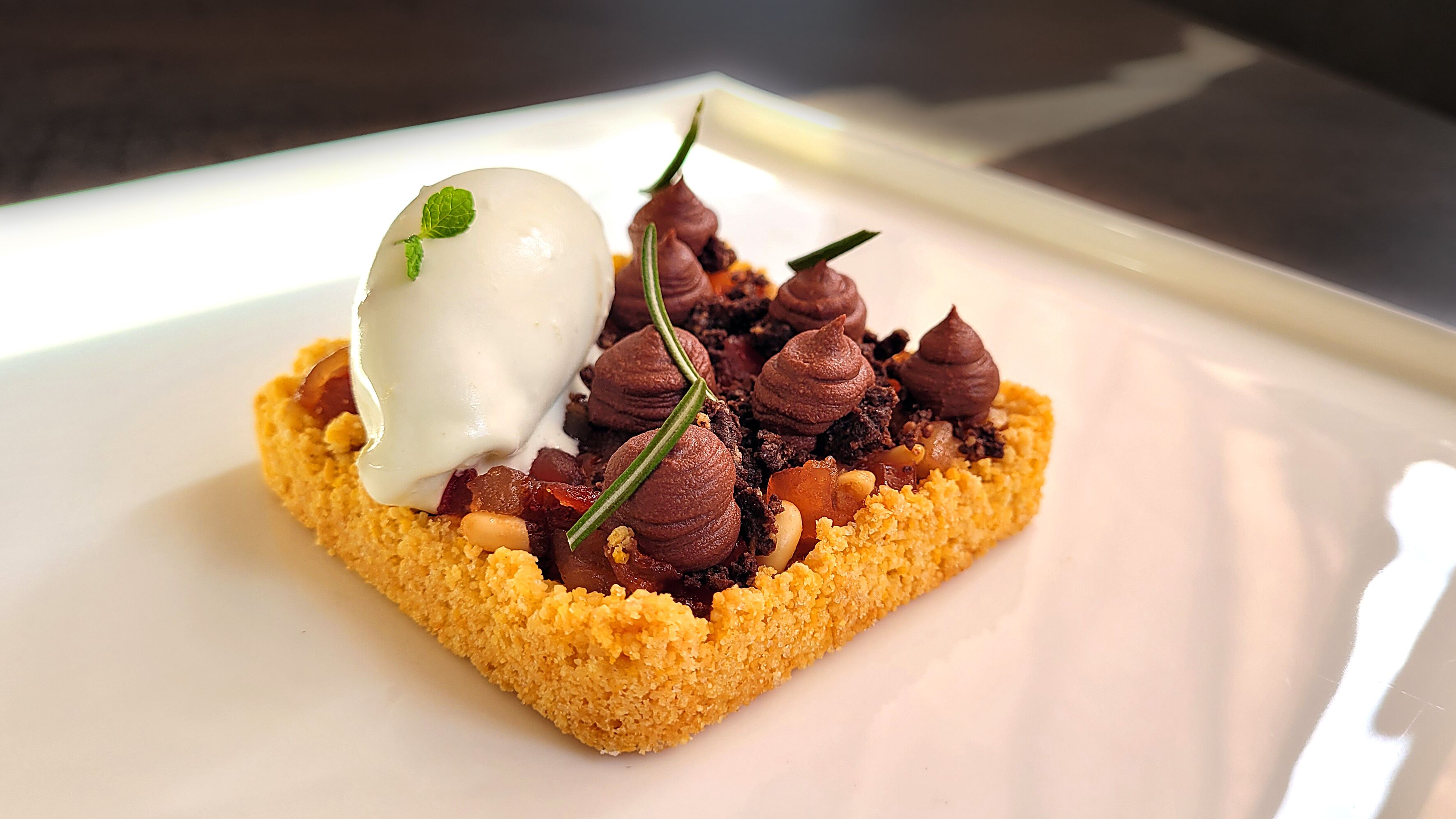 A square-shaped dessert with a creamy topping and chocolate swirls, garnished with a mint leaf, on a white plate.