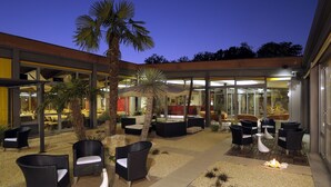 Un espace extérieur aménagé en terrasse, avec des meubles en osier, des palmiers et un coin repas fermé par des parois de verre.
