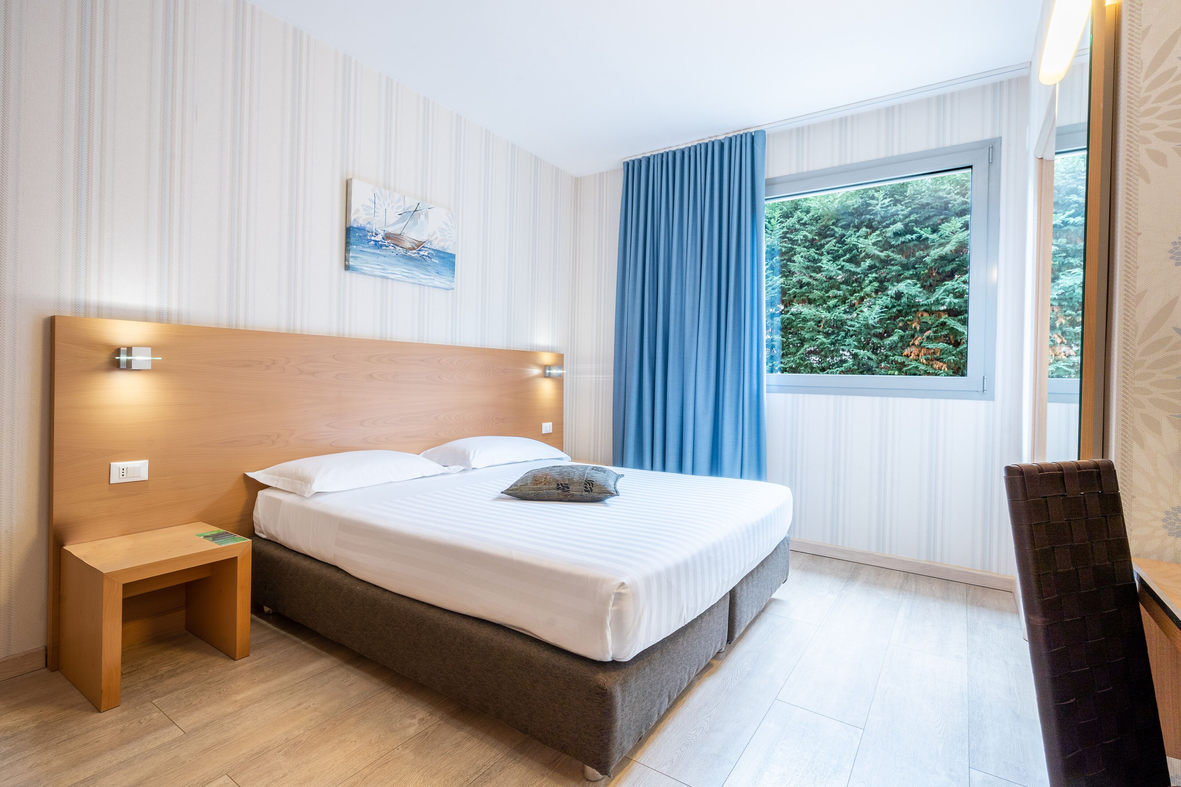 Ett välordnat hotellrum med en stor säng, ett sängbord, en stol och ett fönster med utsikt över grönområden.
