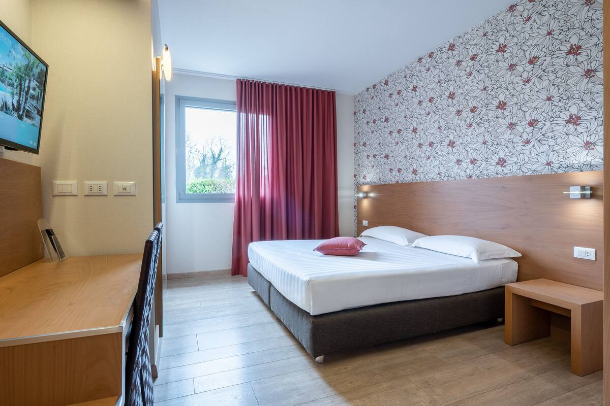 Une chambre d’hôtel comprenant un lit, un bureau, une chaise, une télévision, une fenêtre avec des rideaux et un papier peint à motifs floraux.