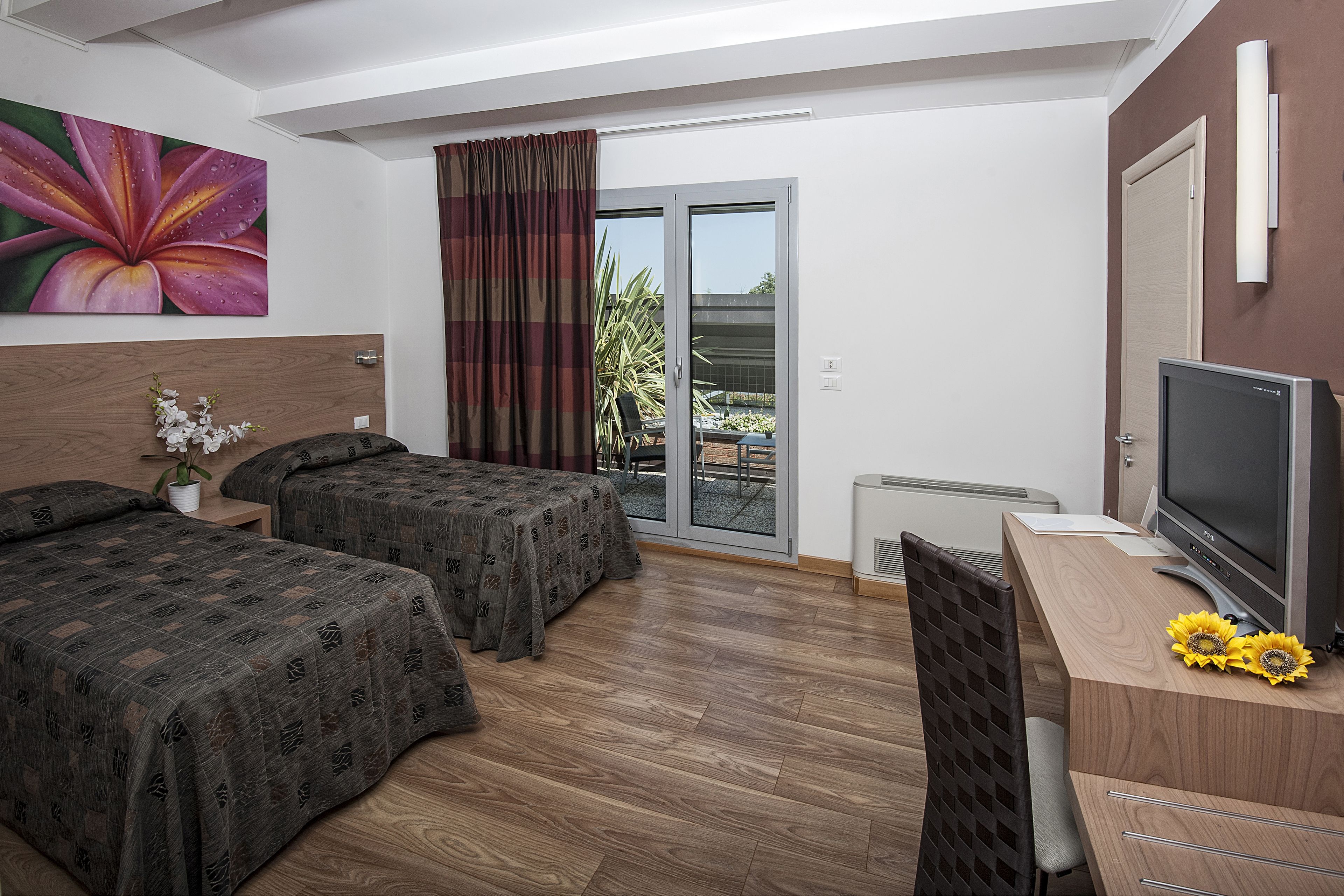 A hotel room with two beds, a desk with a TV, a window with curtains, and a view of the outdoors.