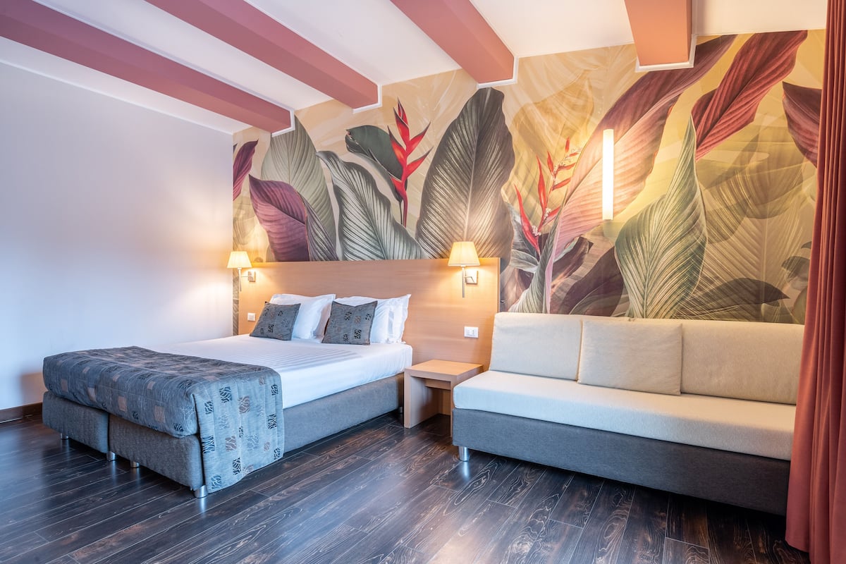 Une chambre d’hôtel moderne, équipée d’un lit, d’un canapé, d’une table de chevet et d’un mur orné d’une fresque représentant des feuilles tropicales.