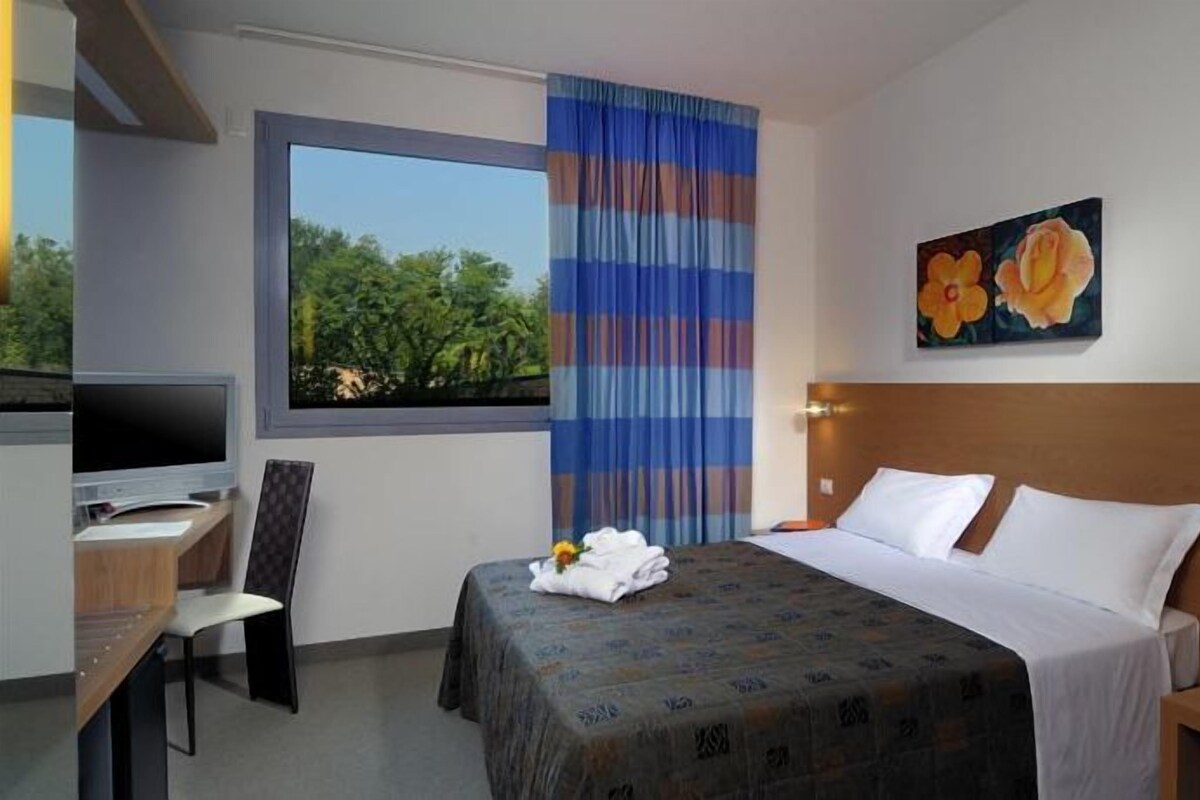 Ein Hotelzimmer mit Bett, Schreibtisch mit Fernseher, Fenster mit Vorhängen und einem Bild an der Wand.