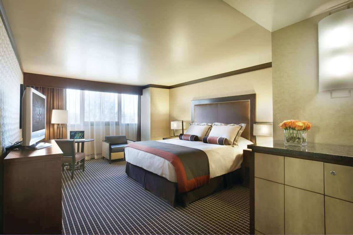 Une chambre d’hôtel avec un grand lit, un bureau avec un ordinateur, une chaise, une petite table avec une lampe et un vase rempli de fleurs sur la commode.