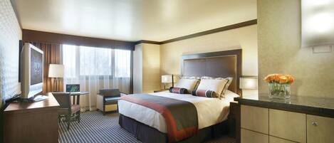 A hotel room with a large bed, a desk with a computer, a chair, a small table with a lamp, and a vase with flowers on the dresser.