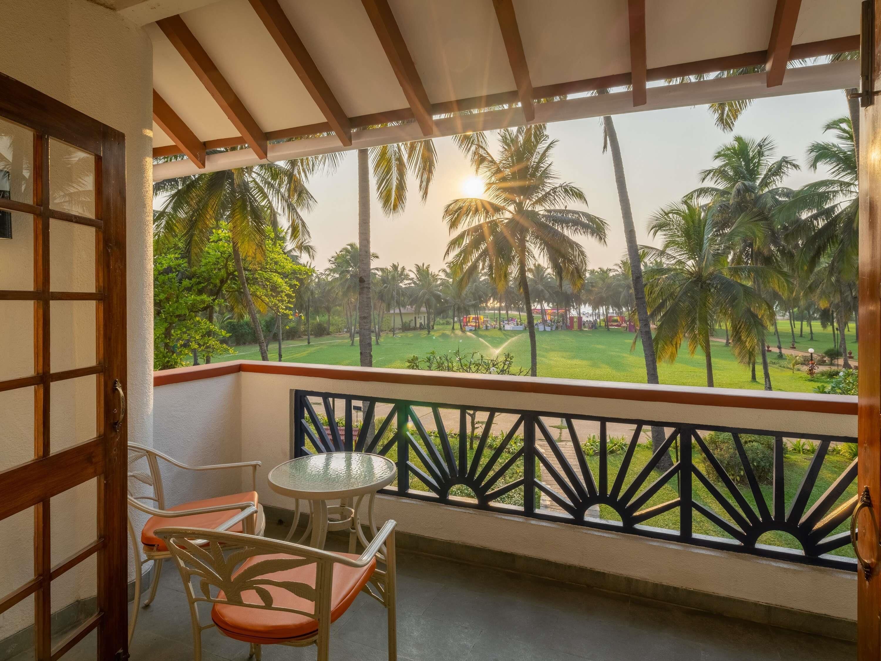A balcony with a view of a lawn and palm trees, a table, and two chairs.