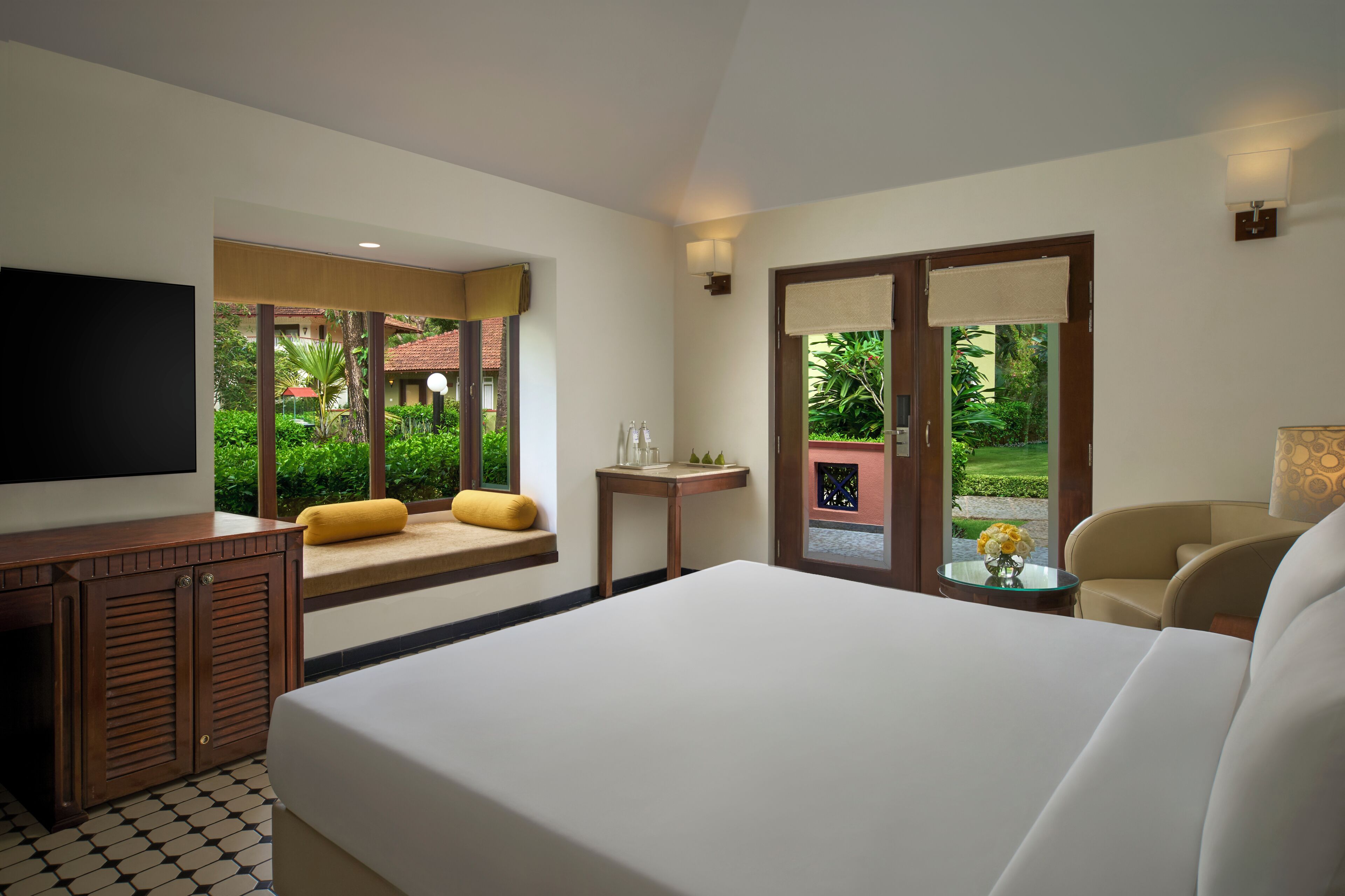 A hotel room with a large bed, a TV, a small table with a vase of flowers, and a seating area with a bench and cushions.