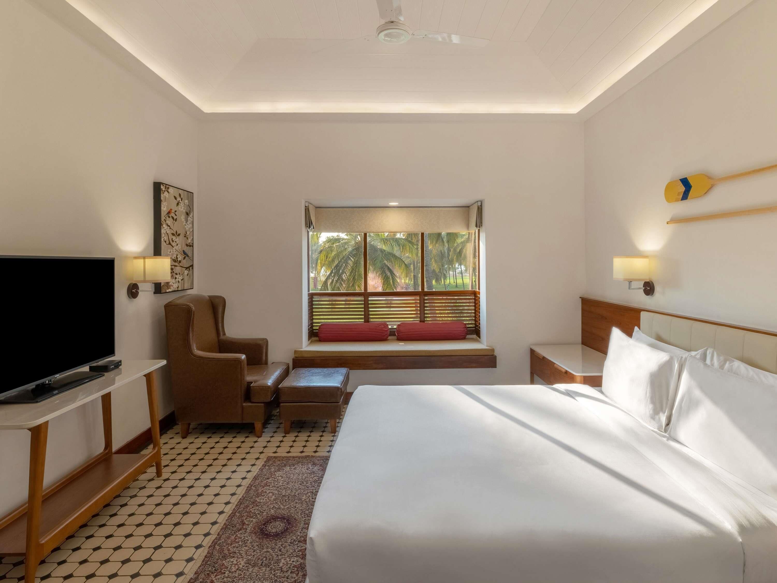 Habitación de hotel con cama, televisión, silla, mesita pequeña, ventana con persianas y ventilador de techo.