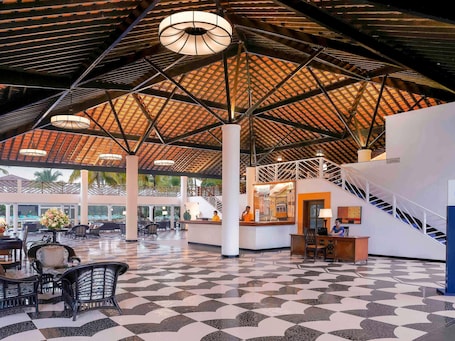 A spacious lobby with a checkered floor, a high ceiling with a patterned design, and a reception desk.. Novotel Goa Dona Sylvia Resort