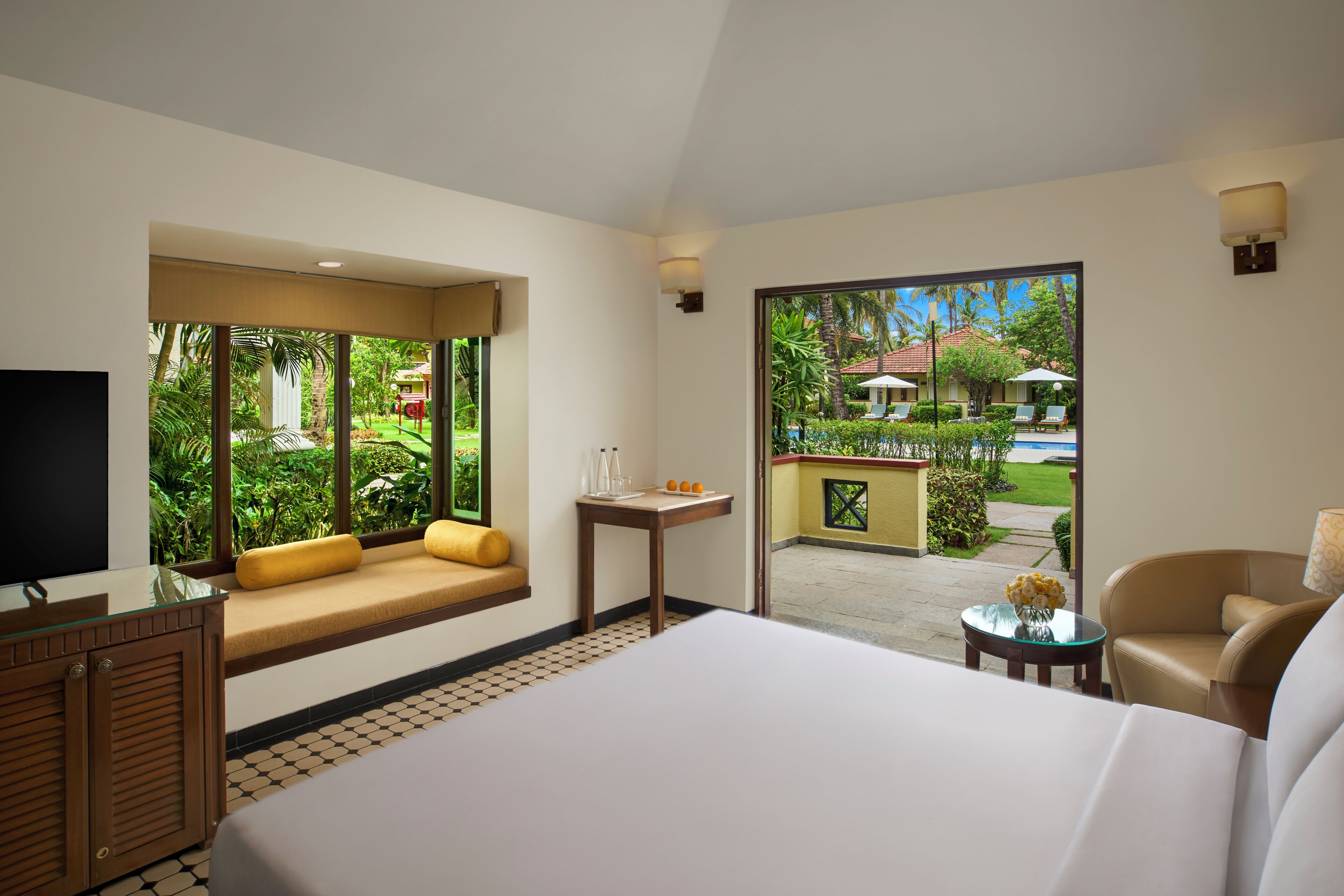 Une chambre d’hôtel dotée d’un grand lit, d’un coin salon avec des coussins, d’une télévision, d’une petite table ornée d’un vase de fleurs, et offrant une vue sur une piscine et la verdure environnante.