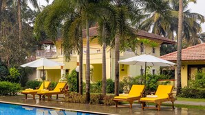 Zona de piscina con sillas y sombrillas amarillas, rodeada de palmeras y un edificio amarillo con tejado de baldosas.