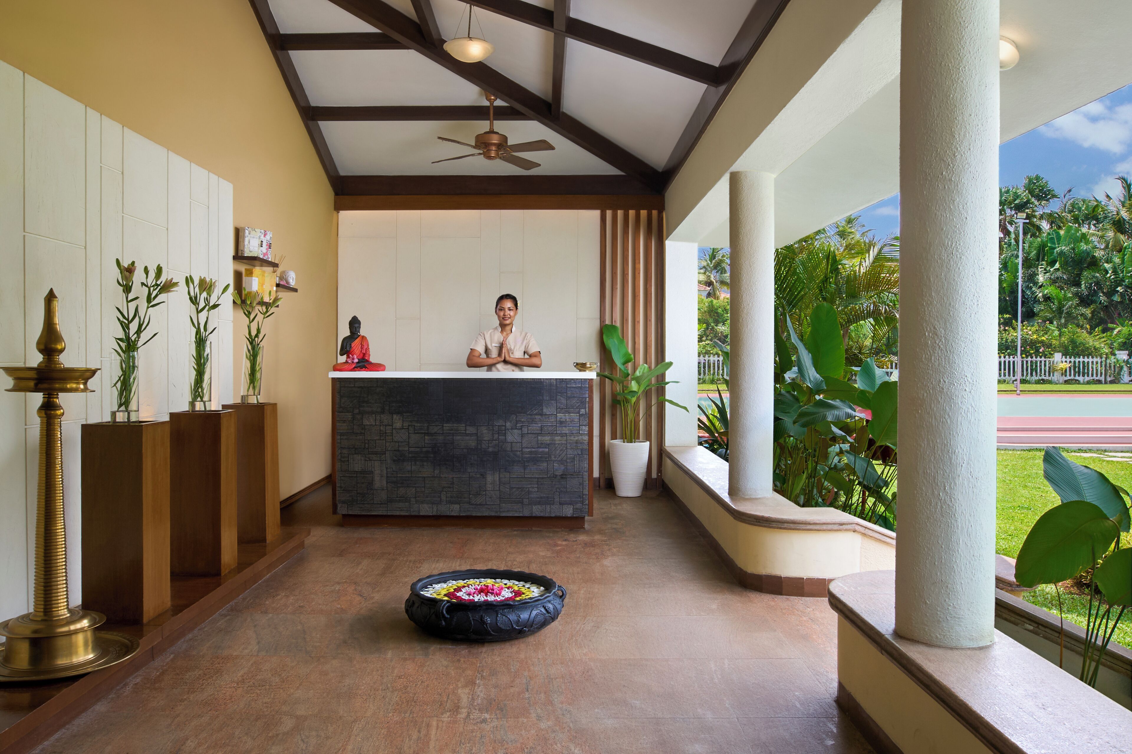 A reception area with a receptionist, a Buddha statue, a ceiling fan, and a view of a garden.
