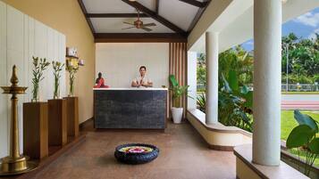 A reception area with a receptionist, a Buddha statue, a ceiling fan, and a view of a garden.