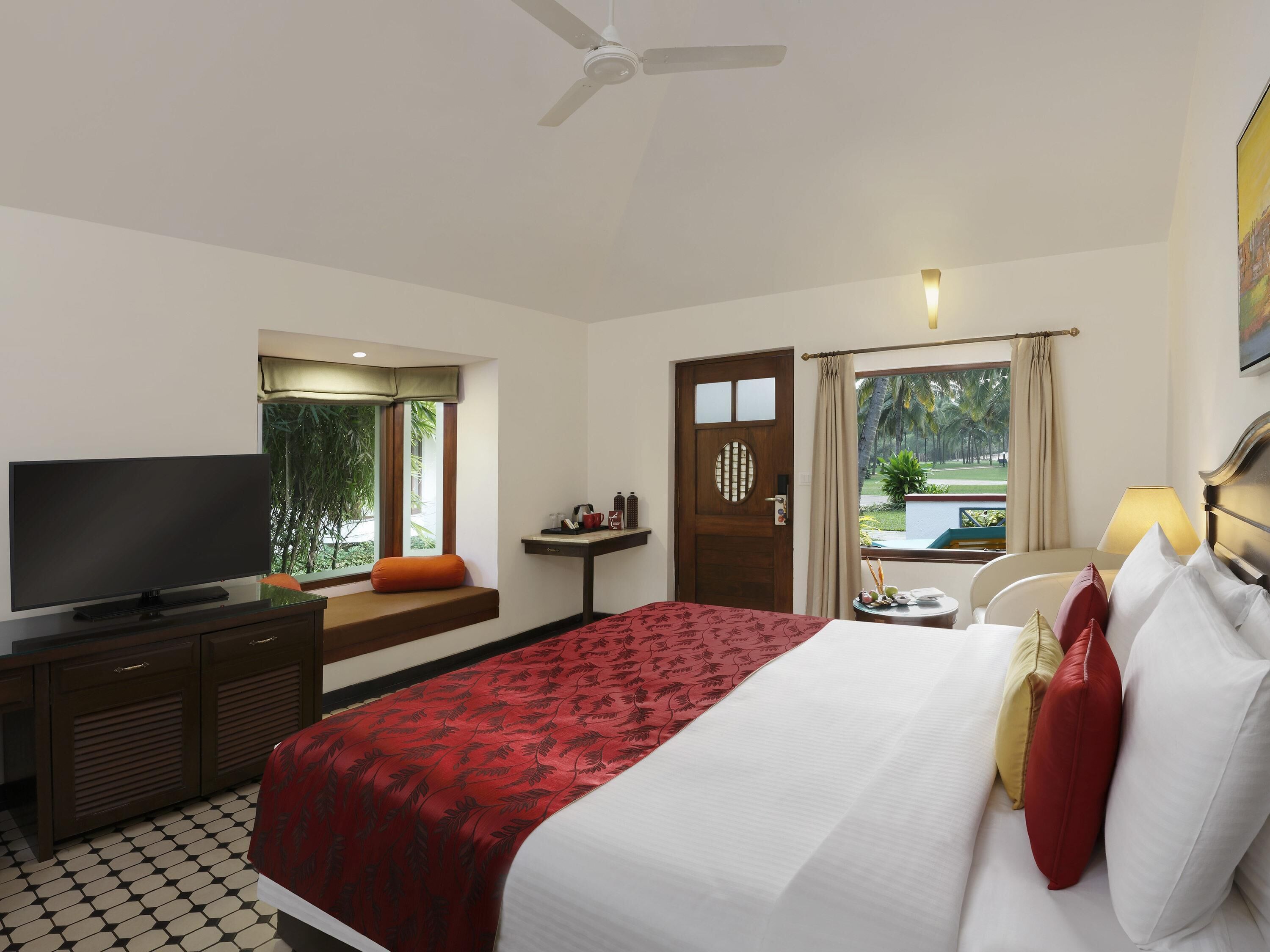 A hotel room with a bed, a TV, a desk, a window with a view of palm trees, and a ceiling fan.
