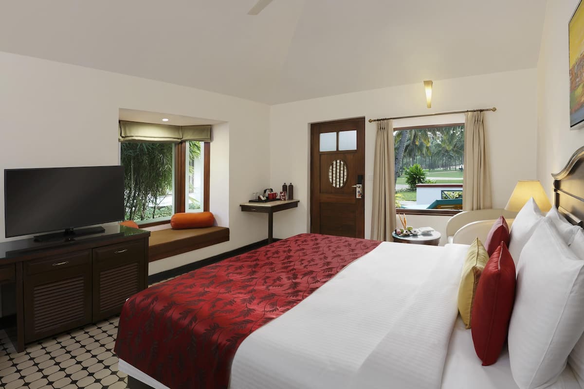 A hotel room with a bed, a TV, a desk, a window with a view of palm trees, and a ceiling fan.
