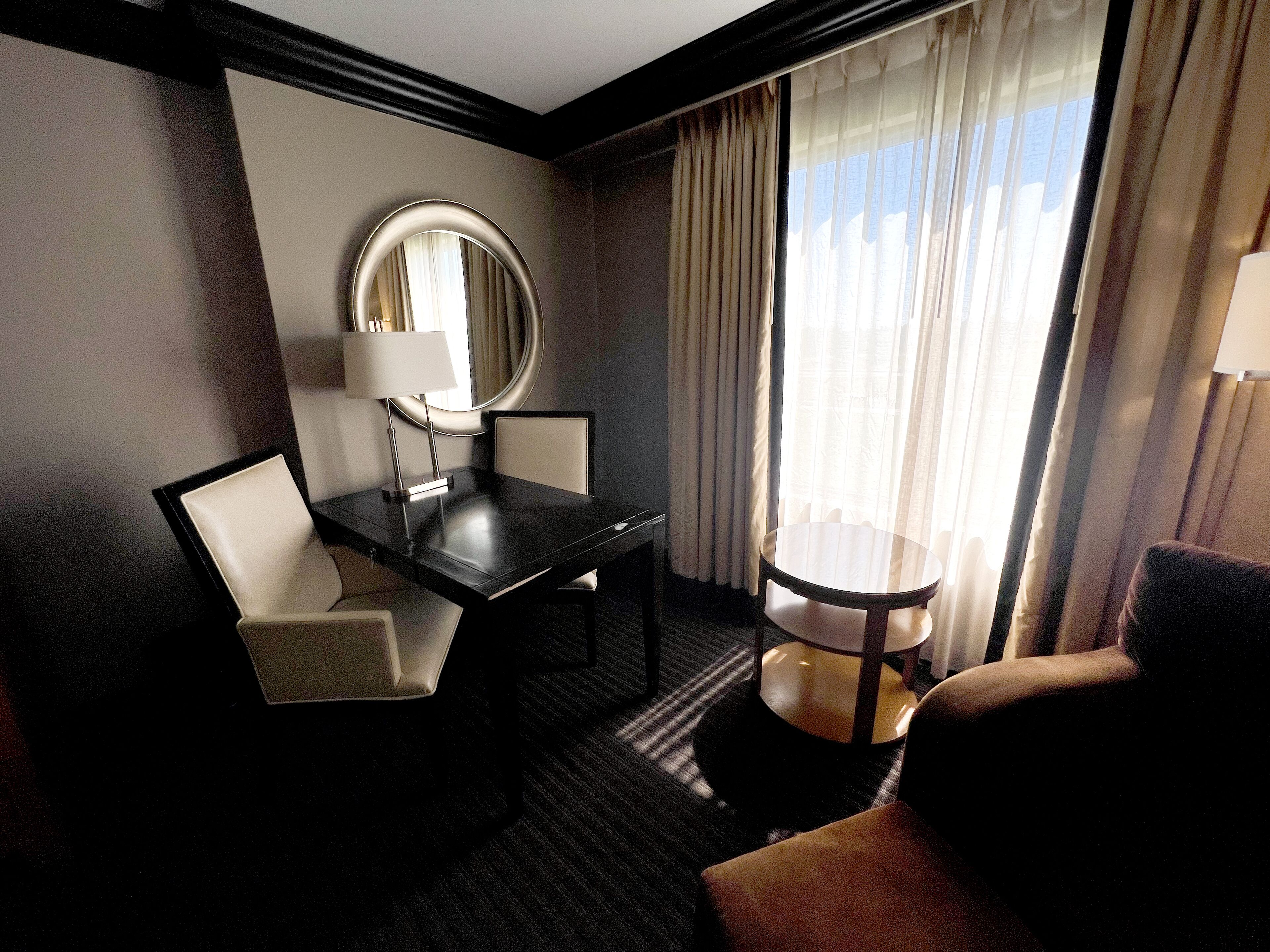 Een hotelkamer met een bureau, twee stoelen, een ronde tafel, een spiegel en uitzicht op het omliggende landschap door dunne gordijnen.
