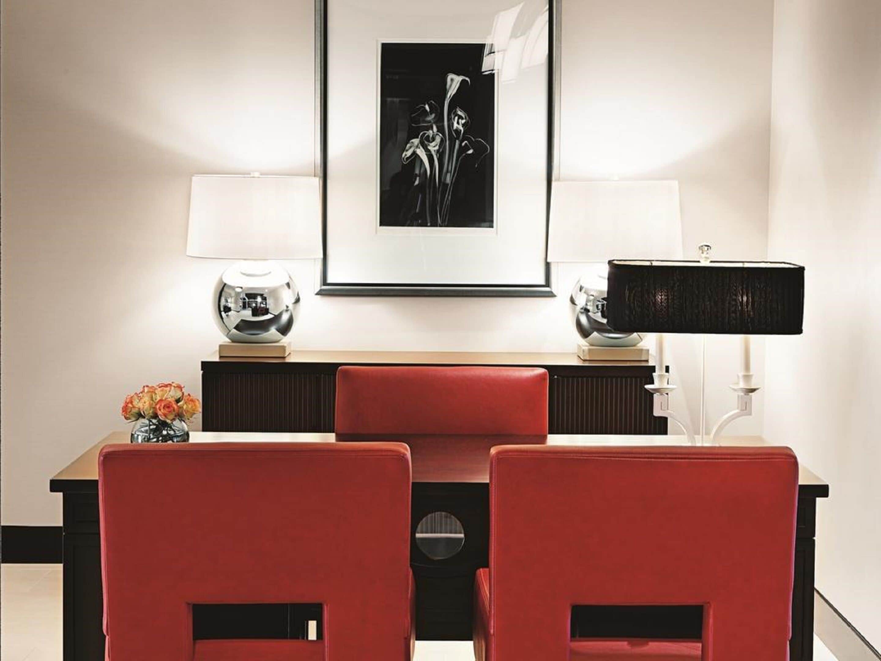 A modern hotel room with a dining area featuring a red upholstered table and chairs, a framed black and white photograph above the table, and two lamps on either side.