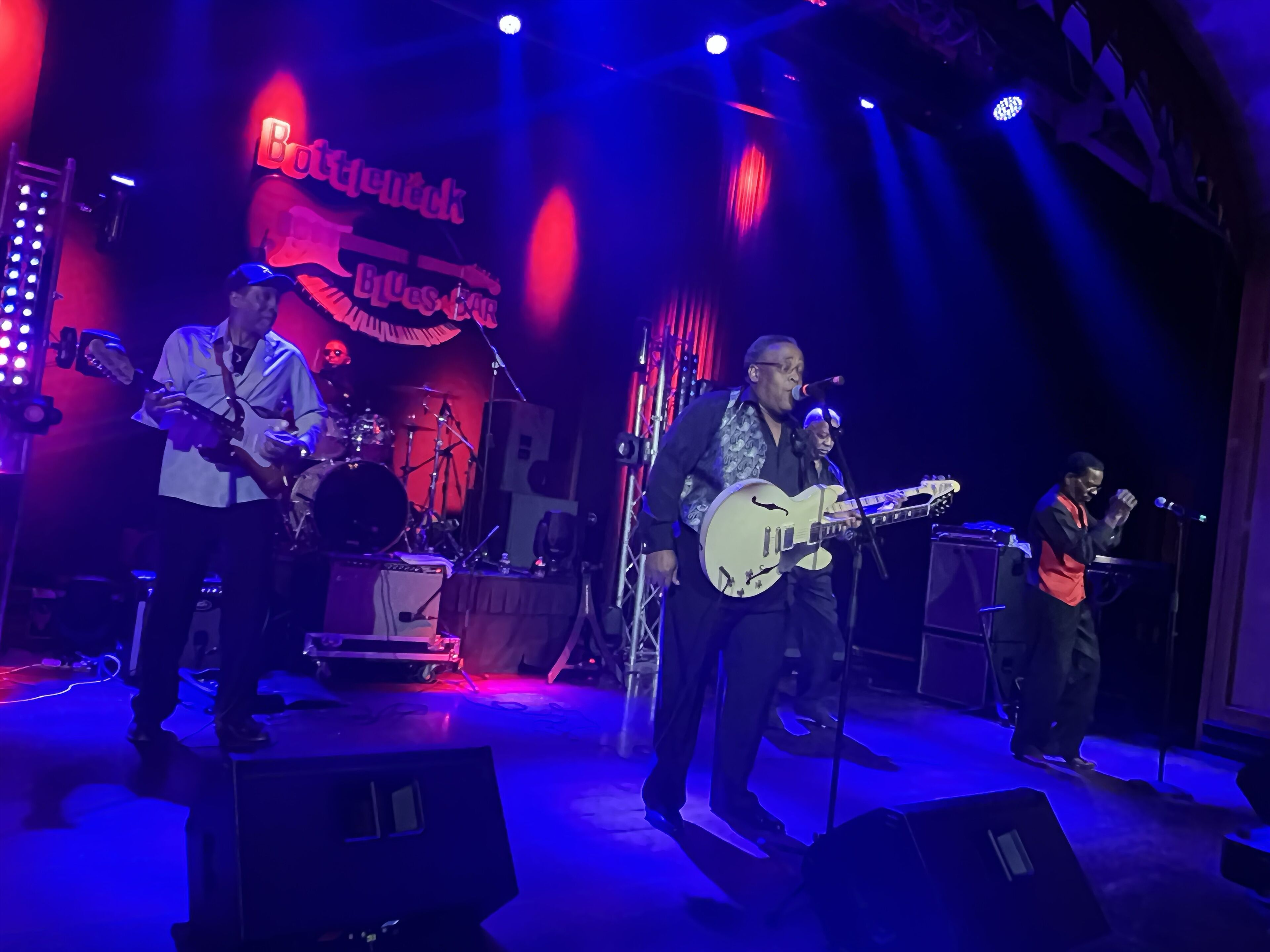 A band performing on a stage with a neon sign reading "Blues Bar" in the background.