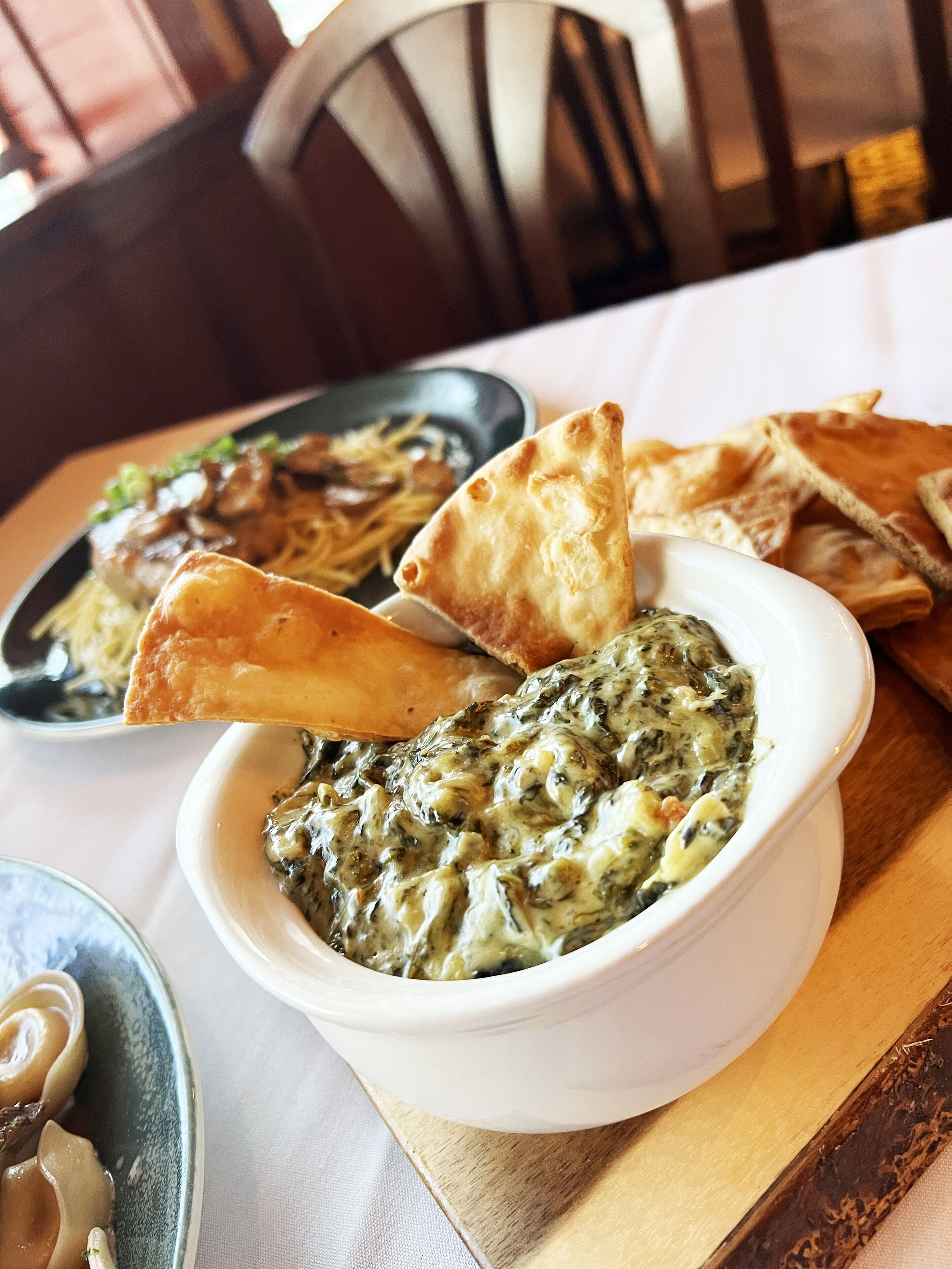 A dining table with a bowl of spinach dip and flatbread, a plate of noodles, and a bowl of soup.