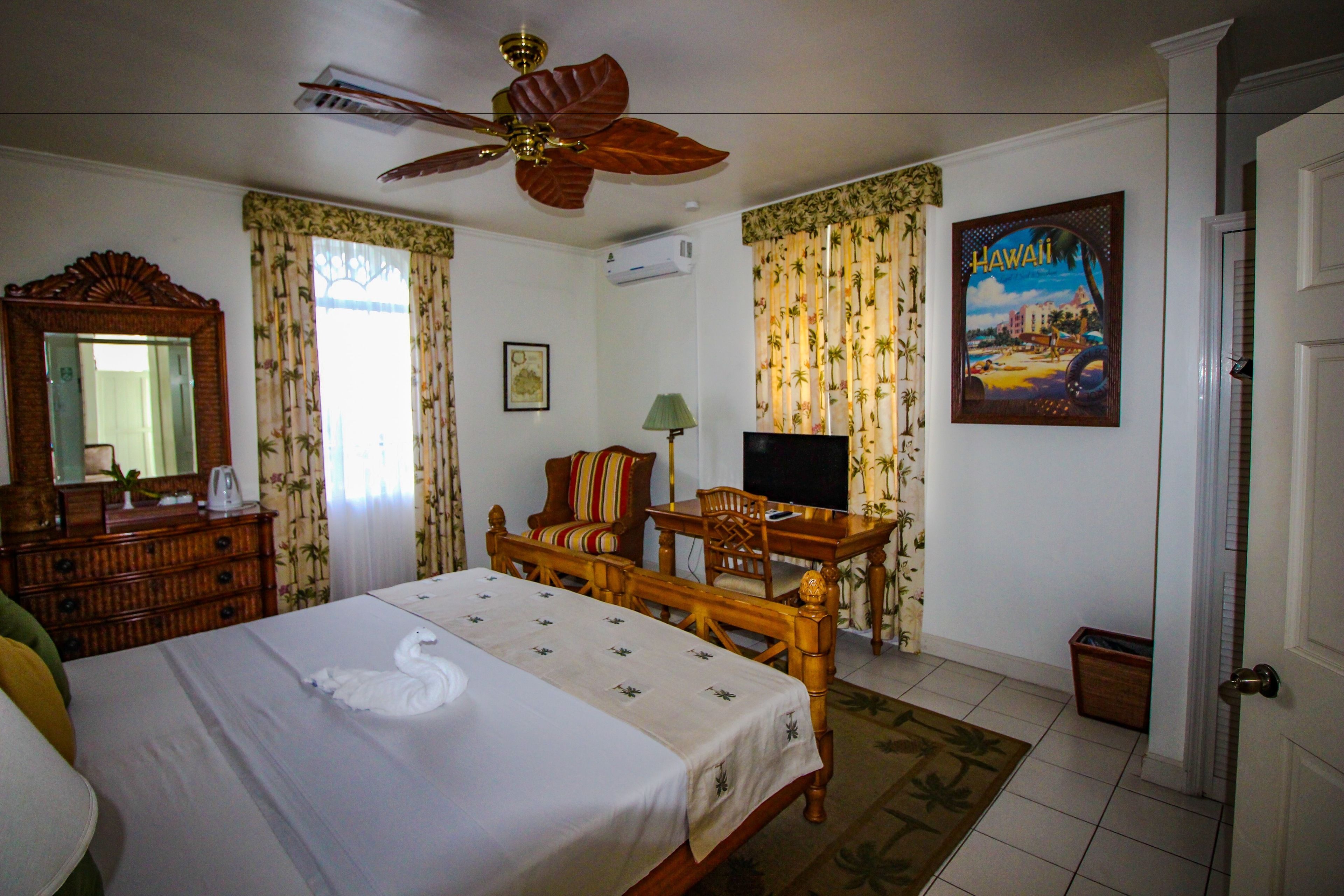 Een hotelkamer met een bed, een houten bureau, een televisie, een plafondventilator en een schilderij van Hawaii.