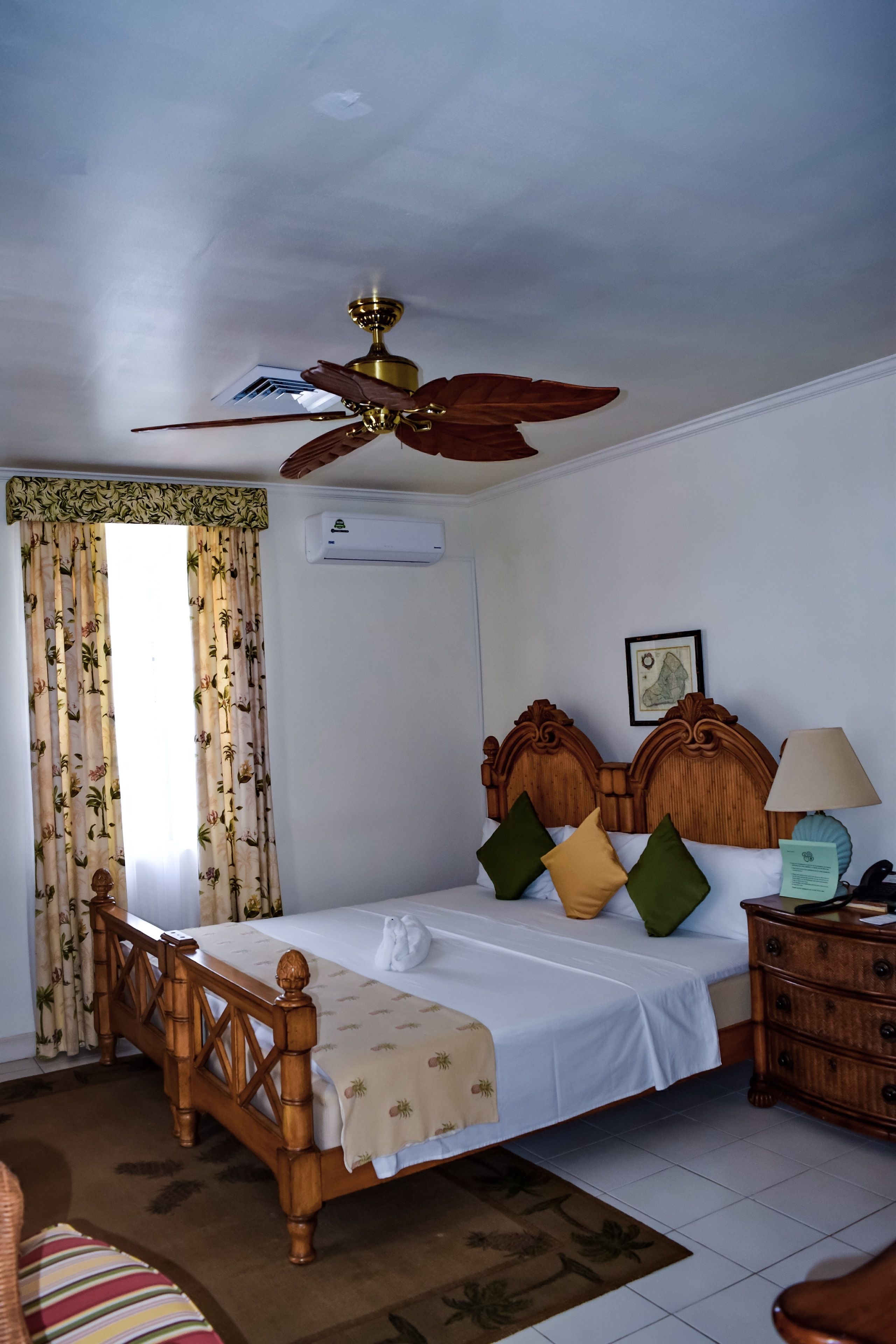 A bedroom with a wooden bed, a ceiling fan, a bedside table with a lamp, and a window with curtains.