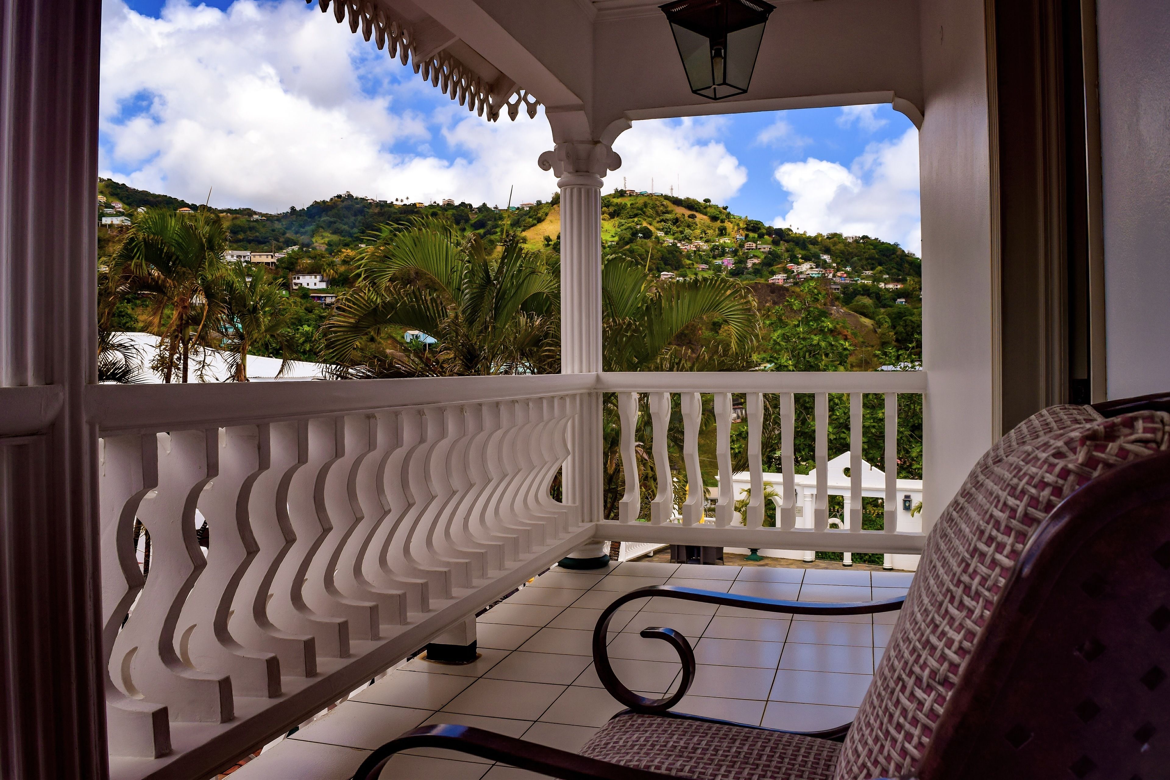 A balcony with a white railing, a wicker chair, and a view of a hillside with houses.
