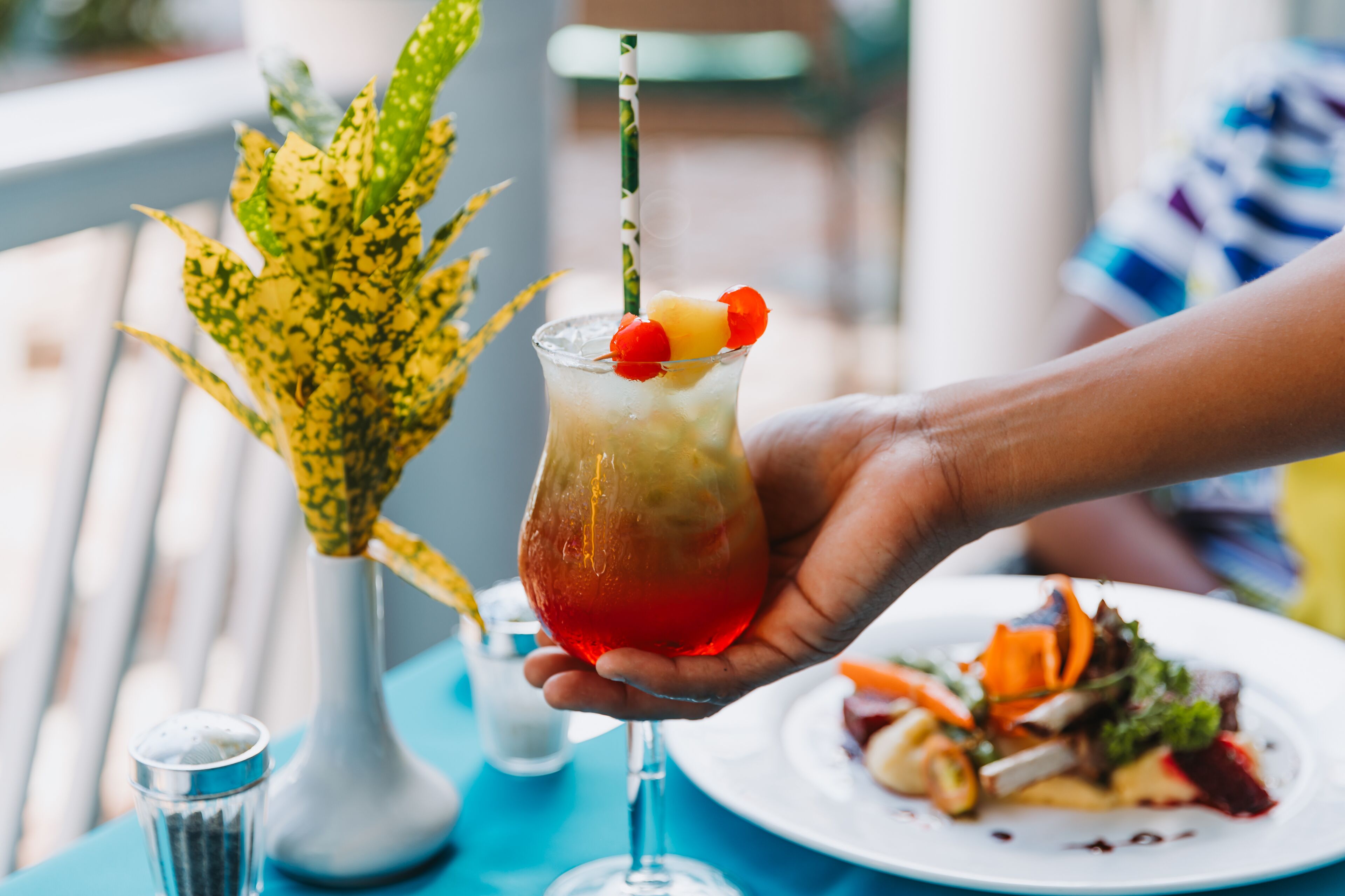 Een hand houdt een cocktail met rietje vast, een bord eten en een vaas met planten op de achtergrond.