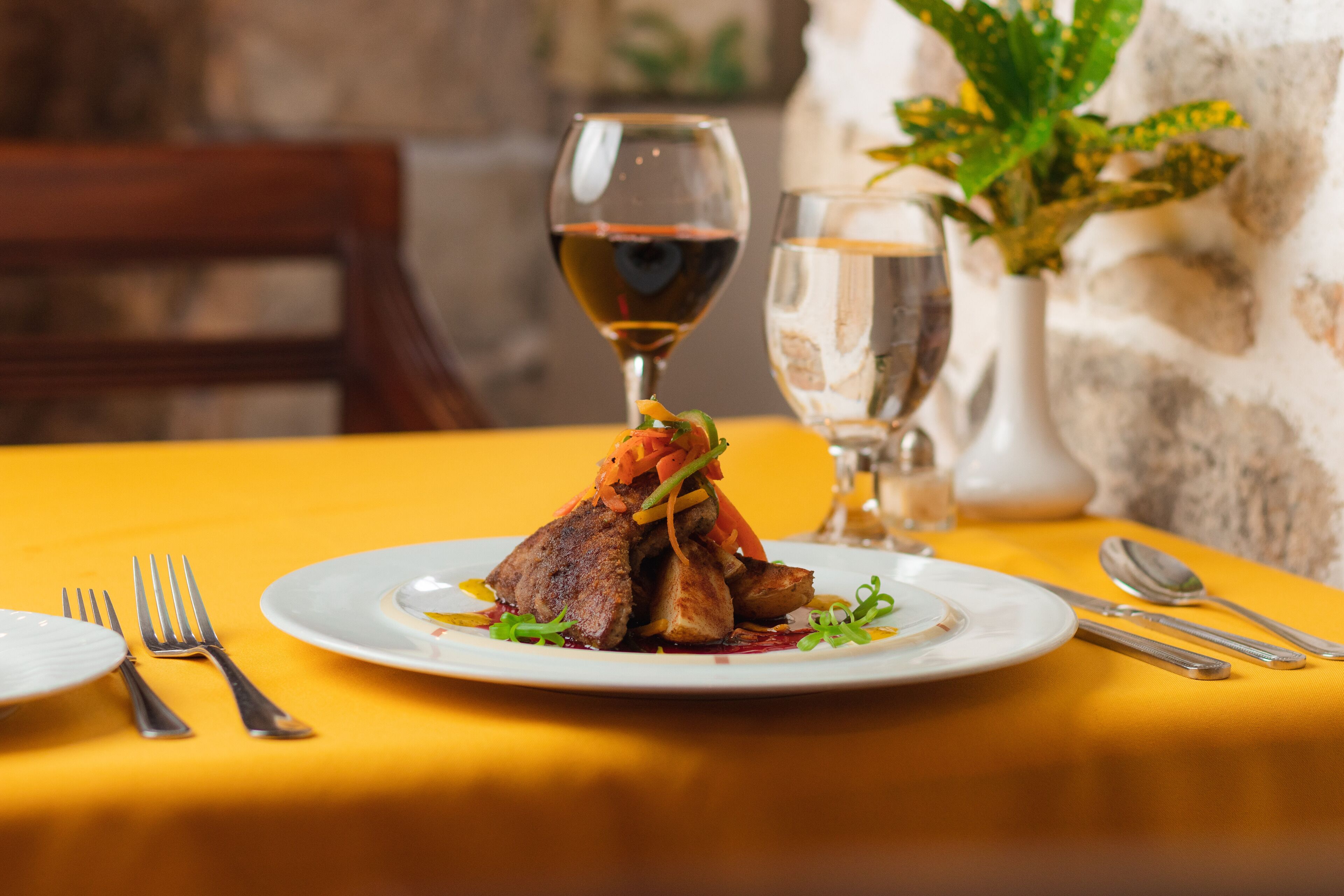 A dining table with a yellow tablecloth, a plate of food, and a glass of red wine.