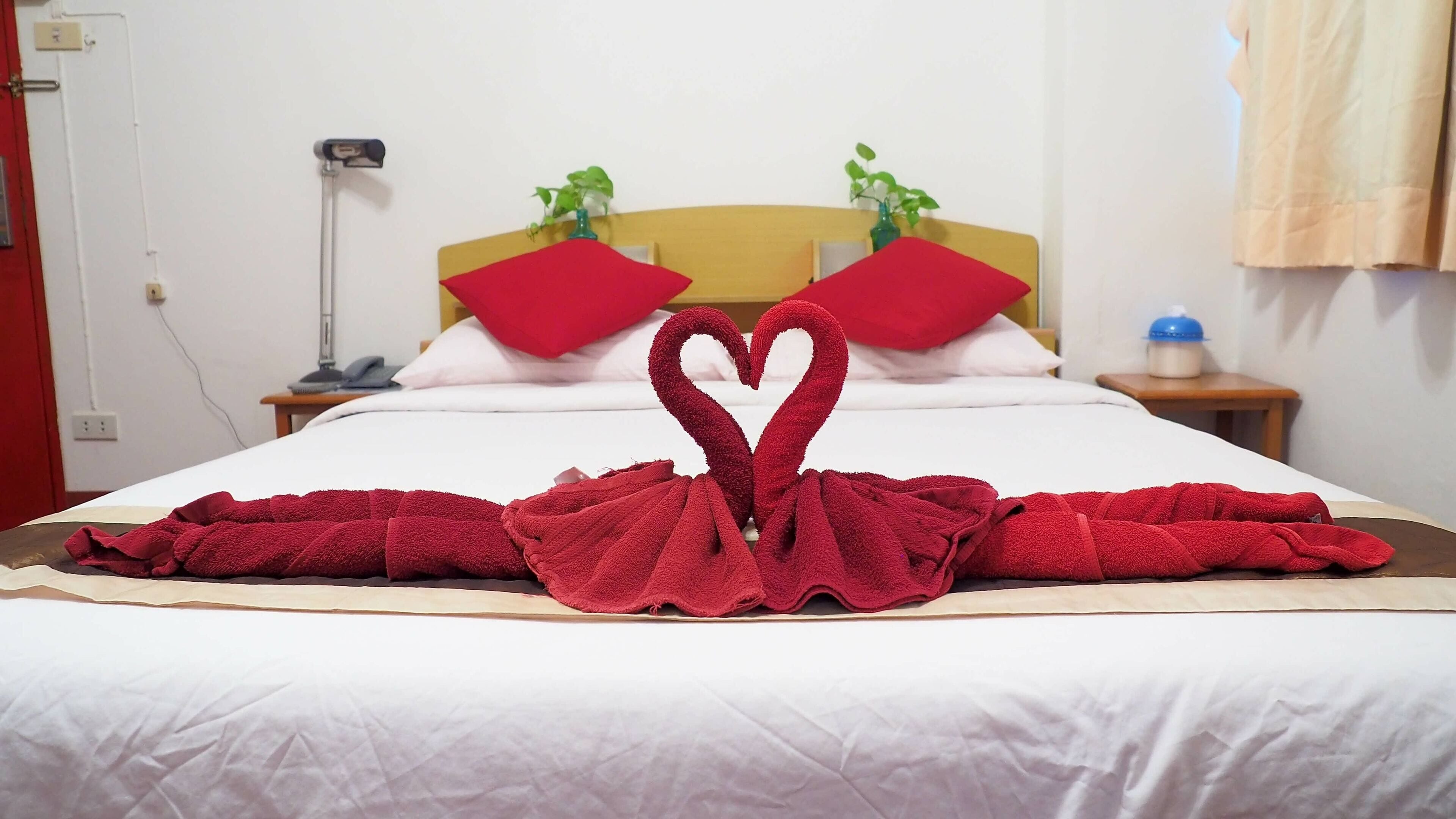 Una cama bien hecha con una disposición de toallas en forma de corazón, un cabecero de madera y una planta en la mesita de noche.