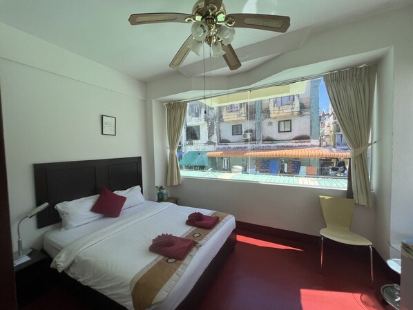 Une chambre d’hôtel avec un lit, une chaise, un ventilateur de plafond et une fenêtre donnant sur les bâtiments environnants.