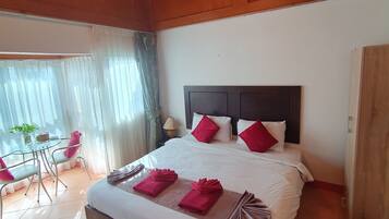 A hotel room with a large bed, a ceiling fan, a dining area with a table and chairs, and a view of the outdoors.