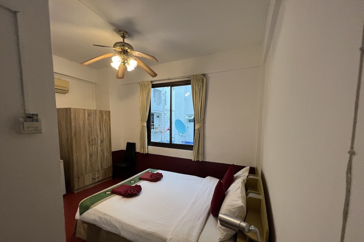 Hotellihuone, jossa on sänky, kattotuuletin, ikkuna verhoilla ja näkymä ulkona oleviin rakennuksiin.