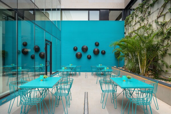 A modern indoor dining area with teal tables and chairs, a blue accent wall, and a planter with greenery.