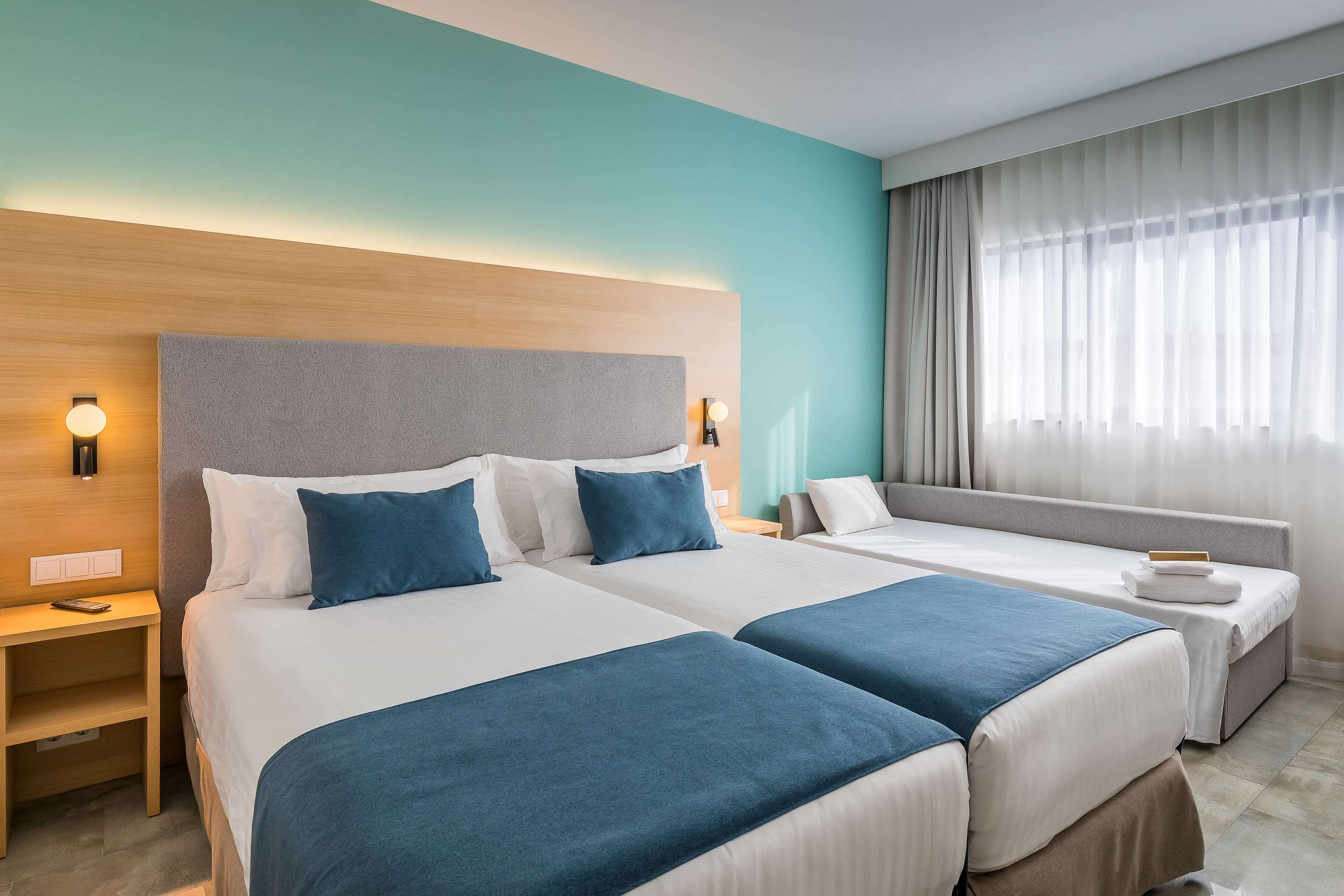 Habitació d'hotel amb dos llits, un sofà, una tauleta de nit i un finestral gran amb cortines.