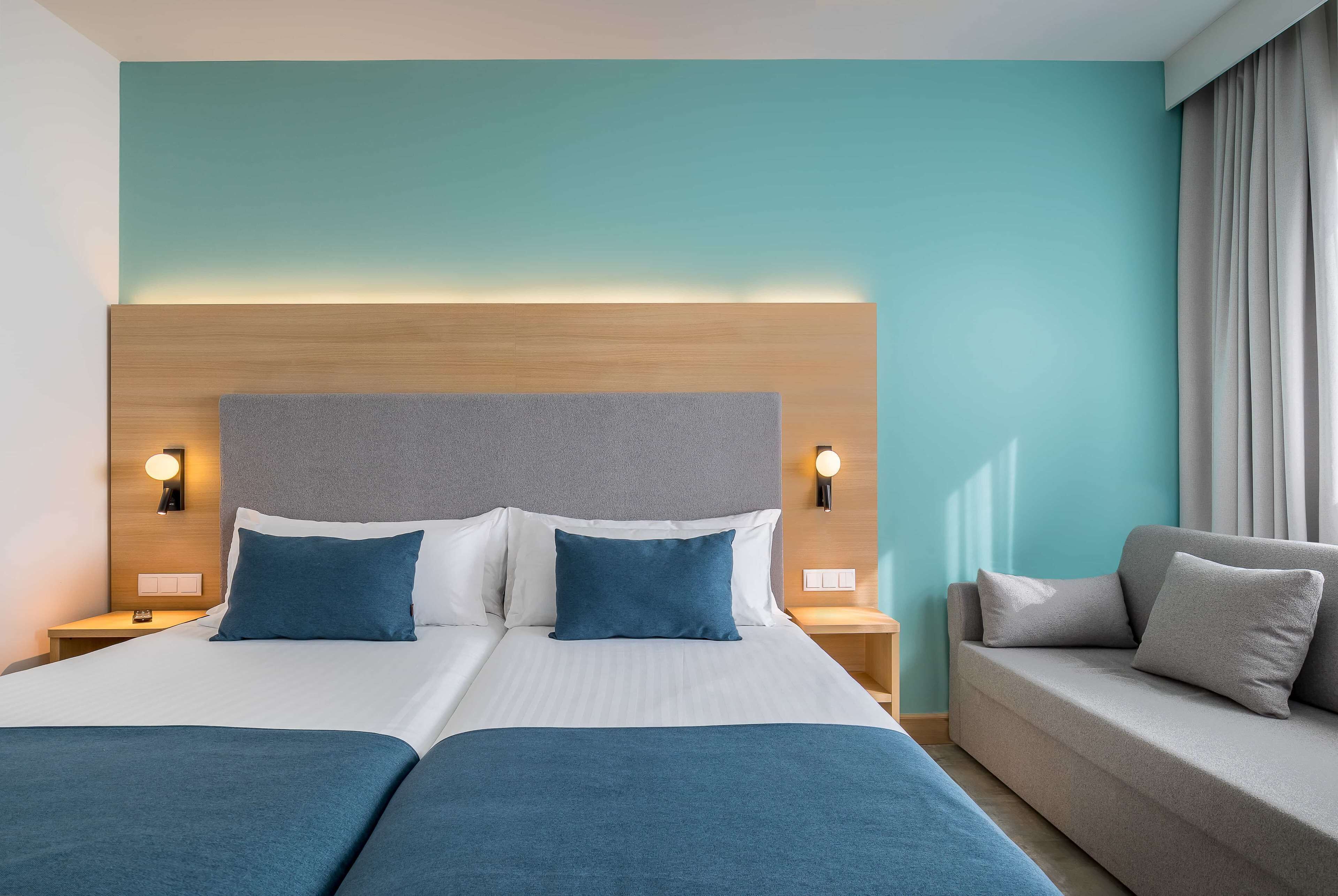 A hotel room with a large bed, two bedside tables, a sofa, and a headboard with integrated lighting.