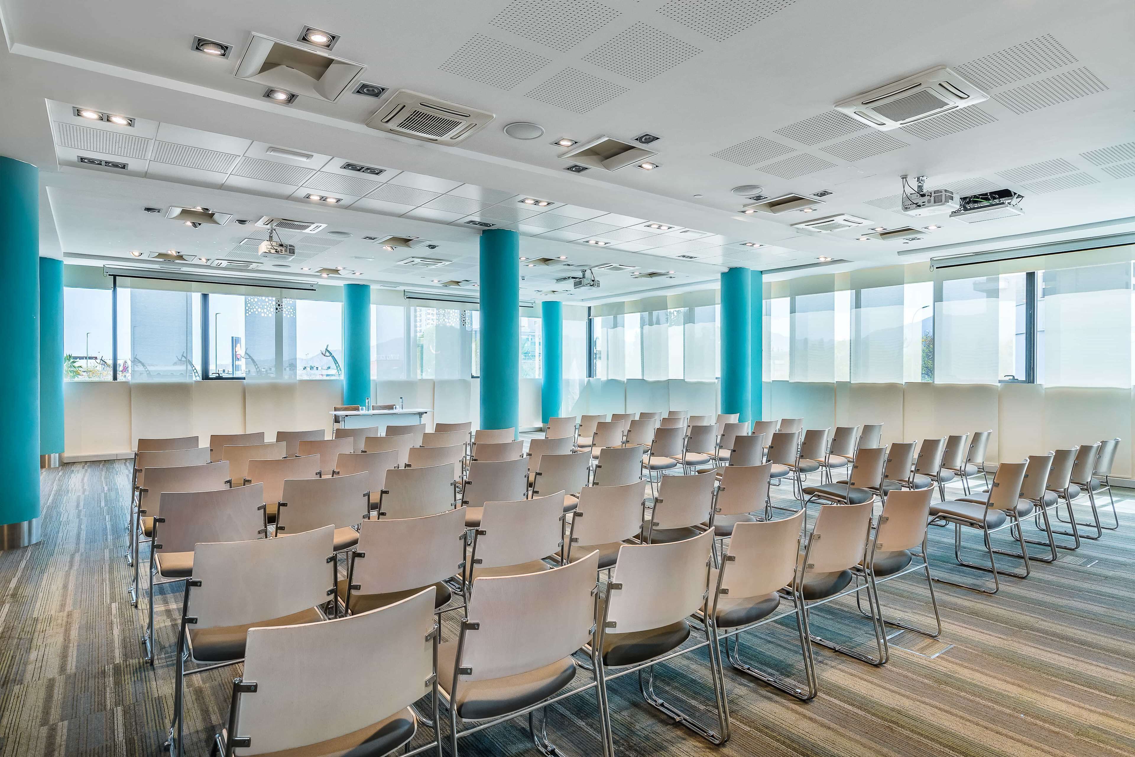 Una sala de conferències amb files de cadires, grans finestres i sortides de ventilació al sostre.