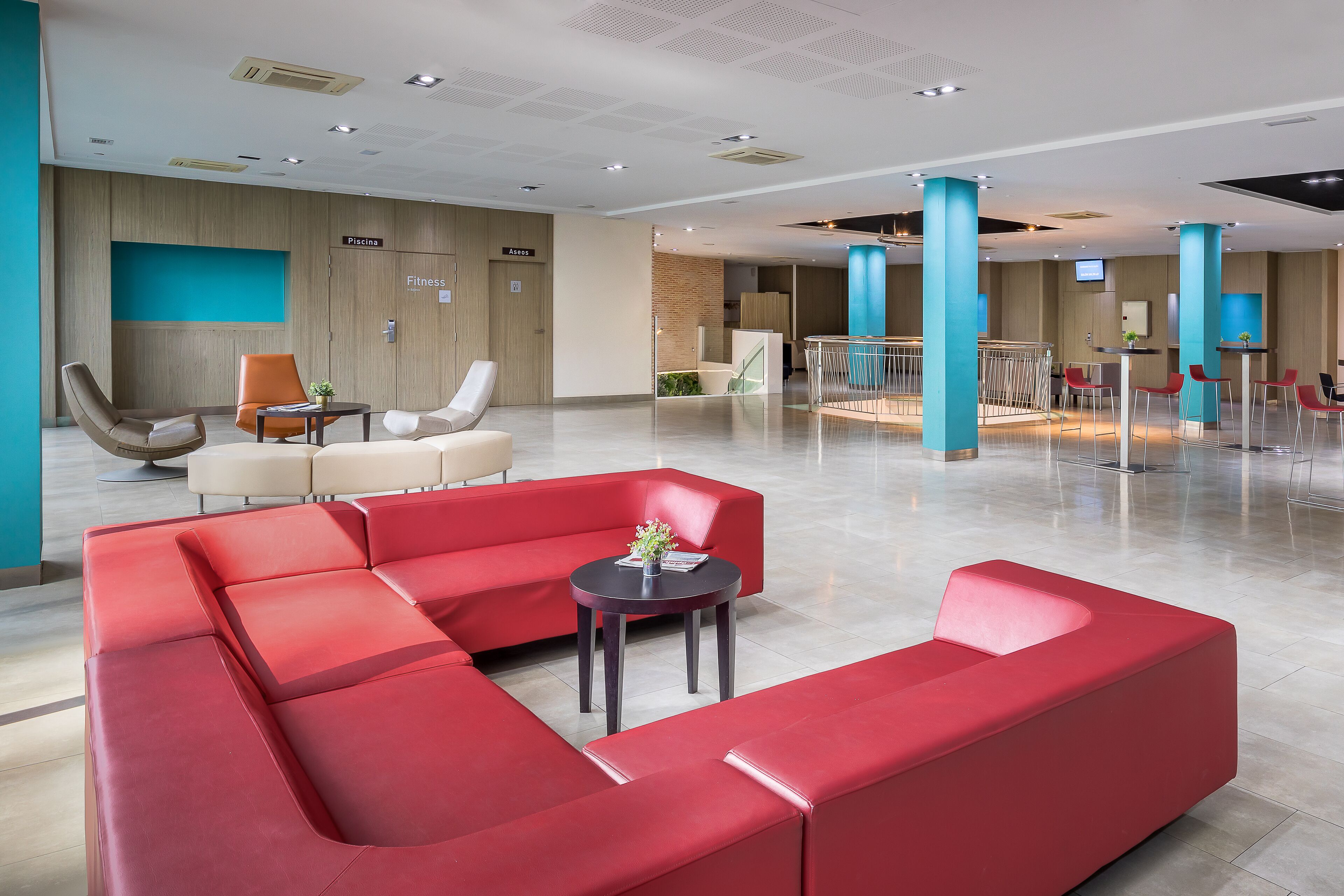 Eine moderne Lobby mit roten Sitzgelegenheiten, einem kleinen Tisch mit Pflanze und einem Schild für den Fitnessbereich.