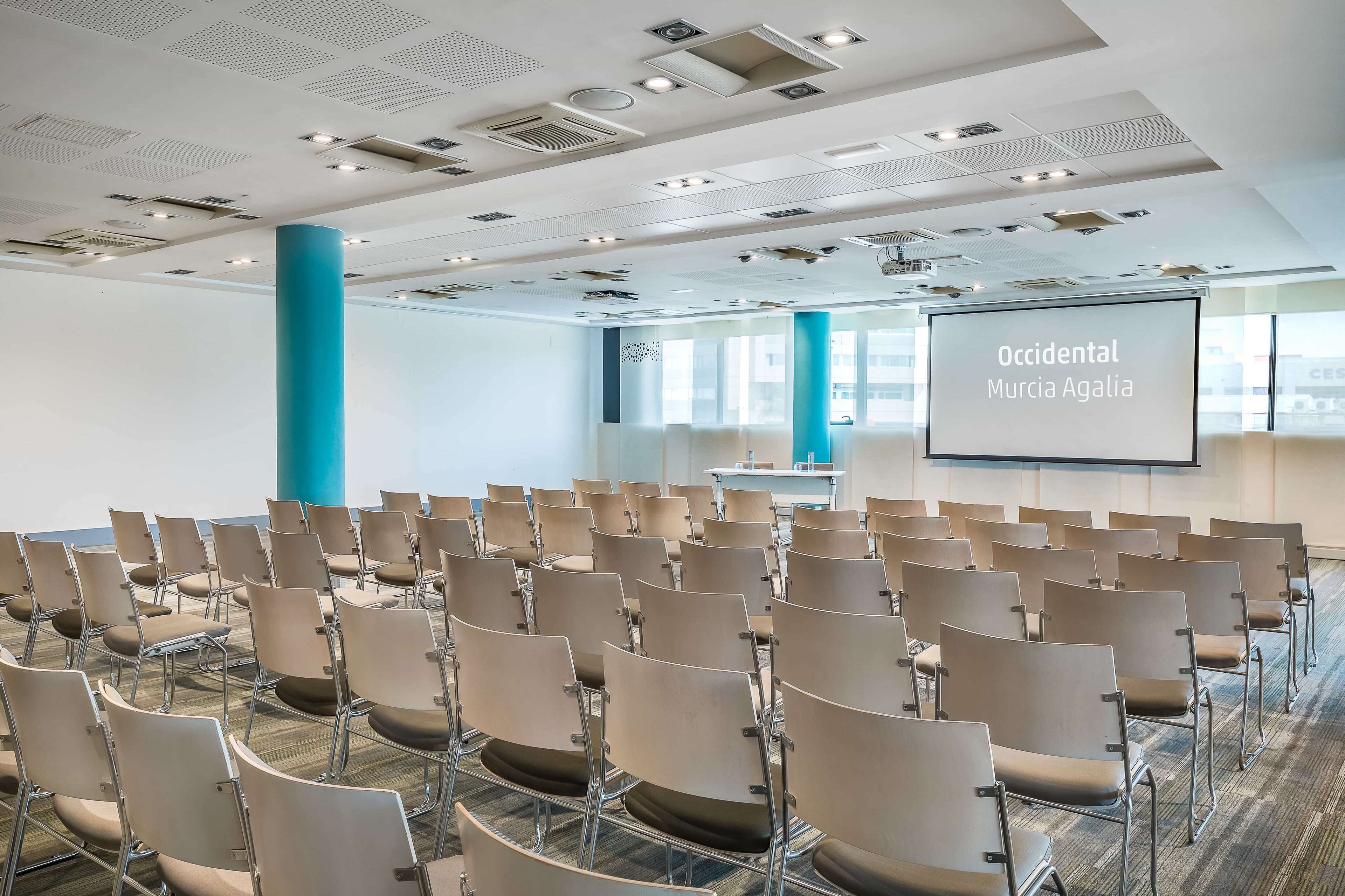 A conference room with rows of chairs, a projector screen displaying "Occidental Murcia Agalia," and a large blue pillar.