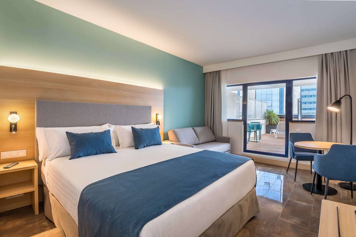 A hotel room with a large bed, a sofa, a desk, and a balcony with a view of the city.