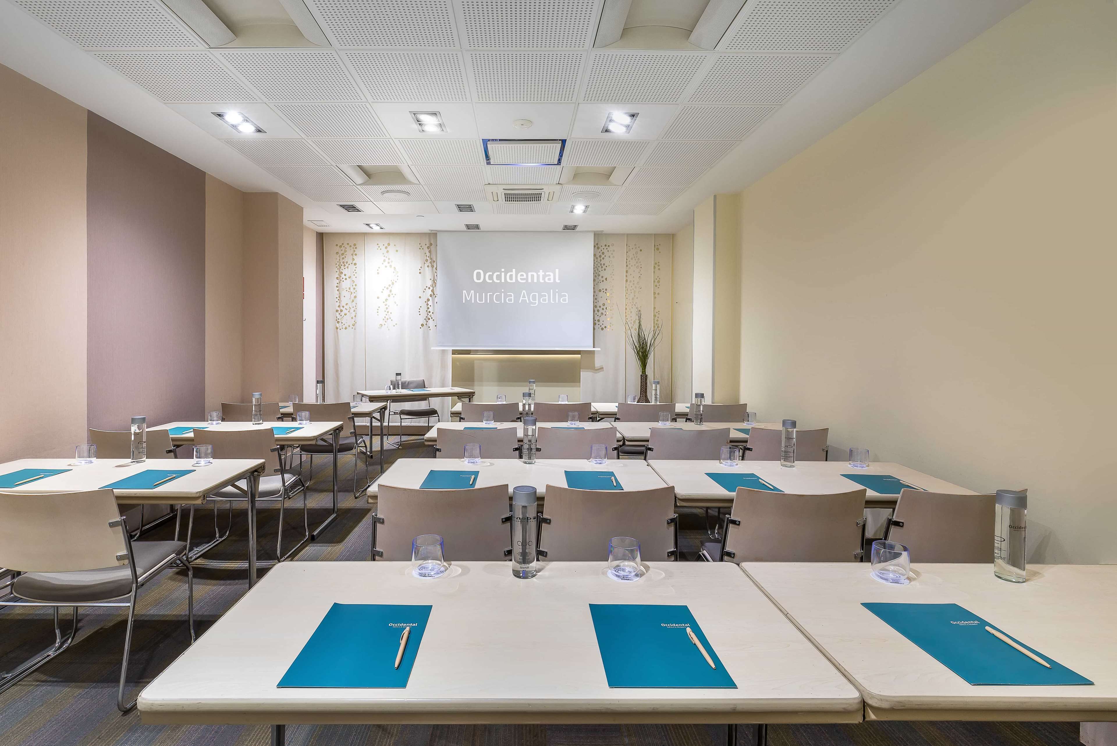 Un salón de conferencias con mesas, sillas y una pantalla para presentaciones.