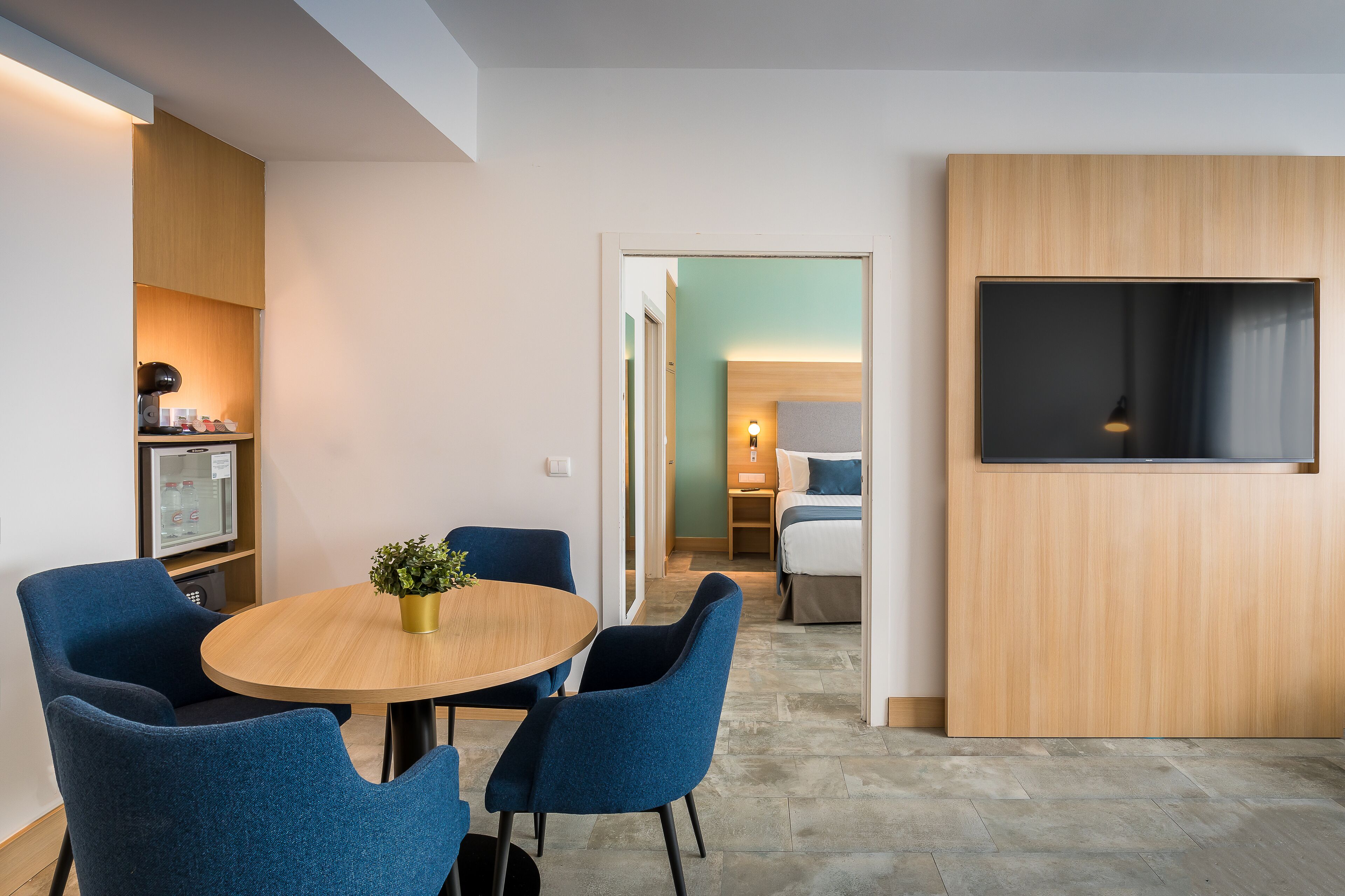 Habitación de hotel moderna con una mesa redonda, sillas azules, una pequeña planta en maceta, un televisor de pantalla plana y una zona de kitchenette.
