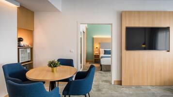 Habitación de hotel moderna con una mesa redonda, sillas azules, una pequeña planta en maceta, un televisor de pantalla plana y una zona de kitchenette.