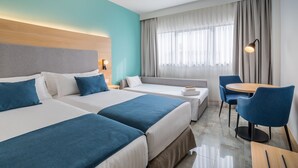 A hotel room with two beds, a sofa, a dining table with chairs, a lamp, and a large window with curtains.