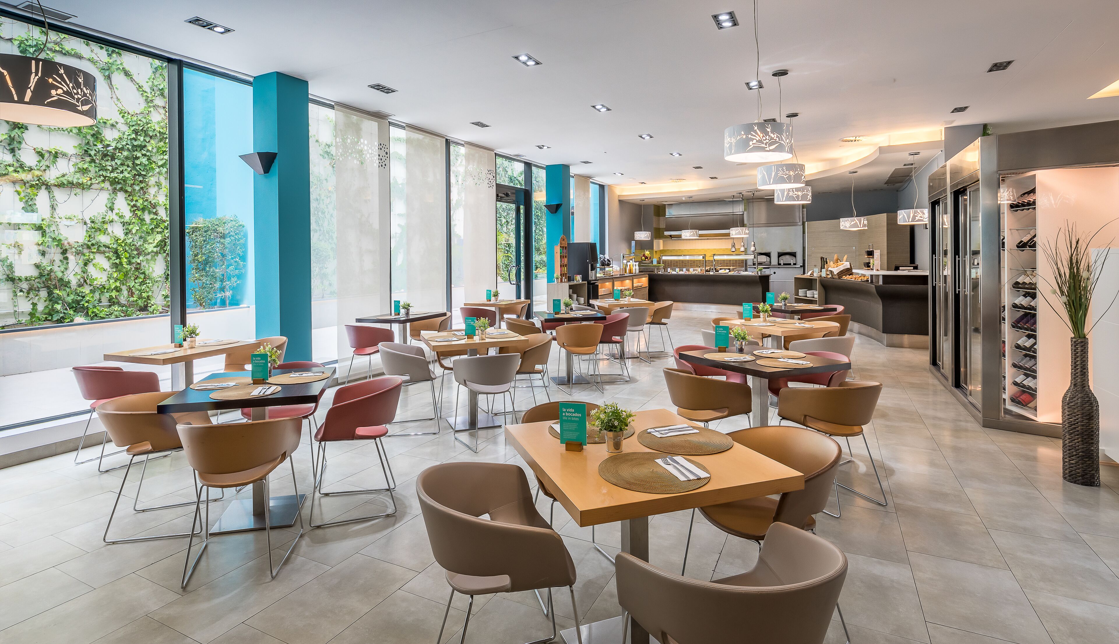 A modern restaurant with multiple tables and chairs, large windows, and a visible kitchen area.