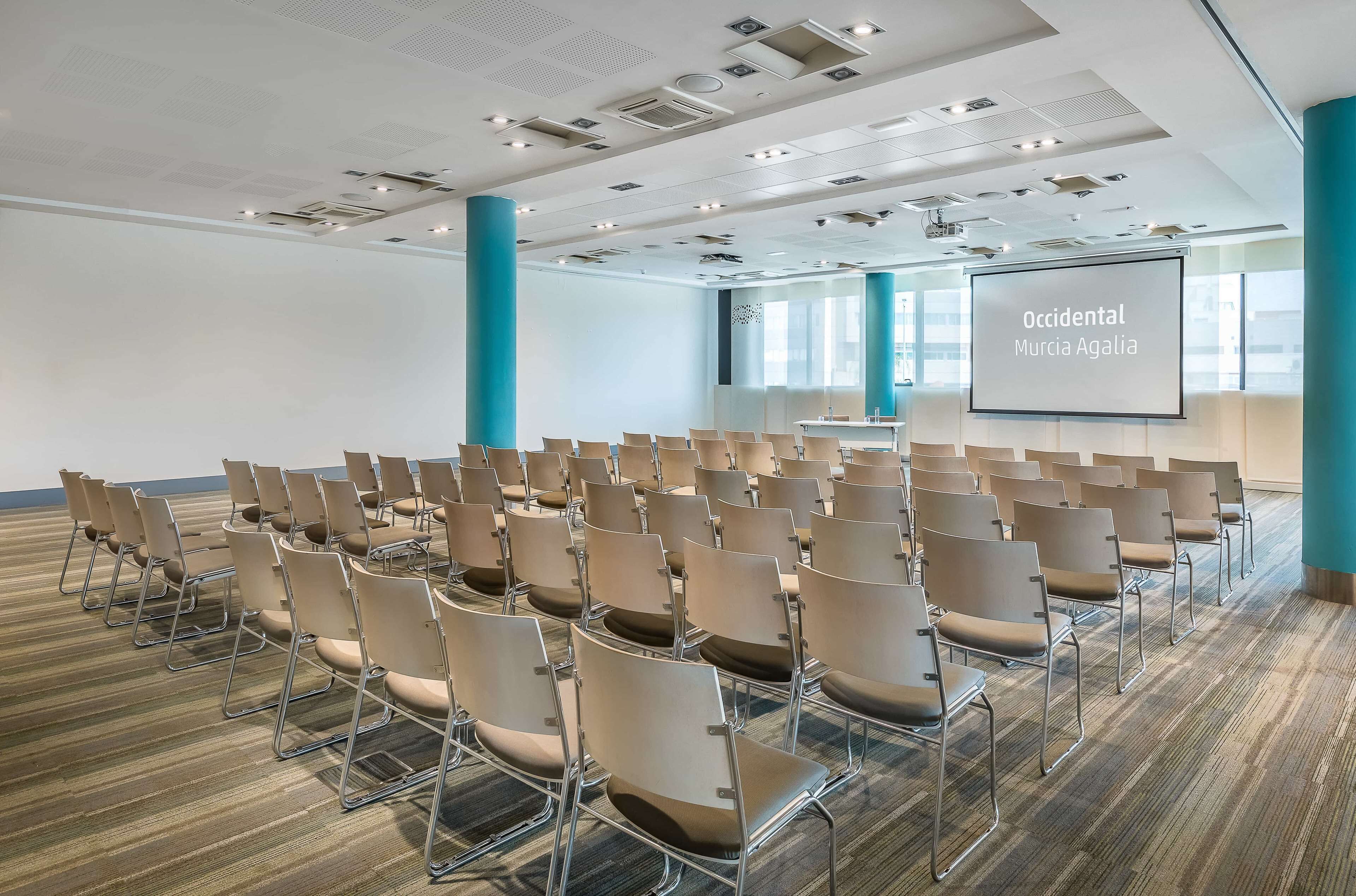 Une salle de conférence avec des rangées de chaises, un écran de projection affichant le texte « Occidental Murcia Agalia », et une grande fenêtre laissant entrer la lumière naturelle.